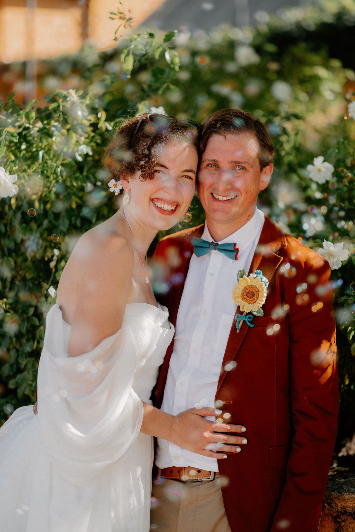 Happy couple smiles in their wedding attire beneath blooming white flowers and greenery on a sunny day.