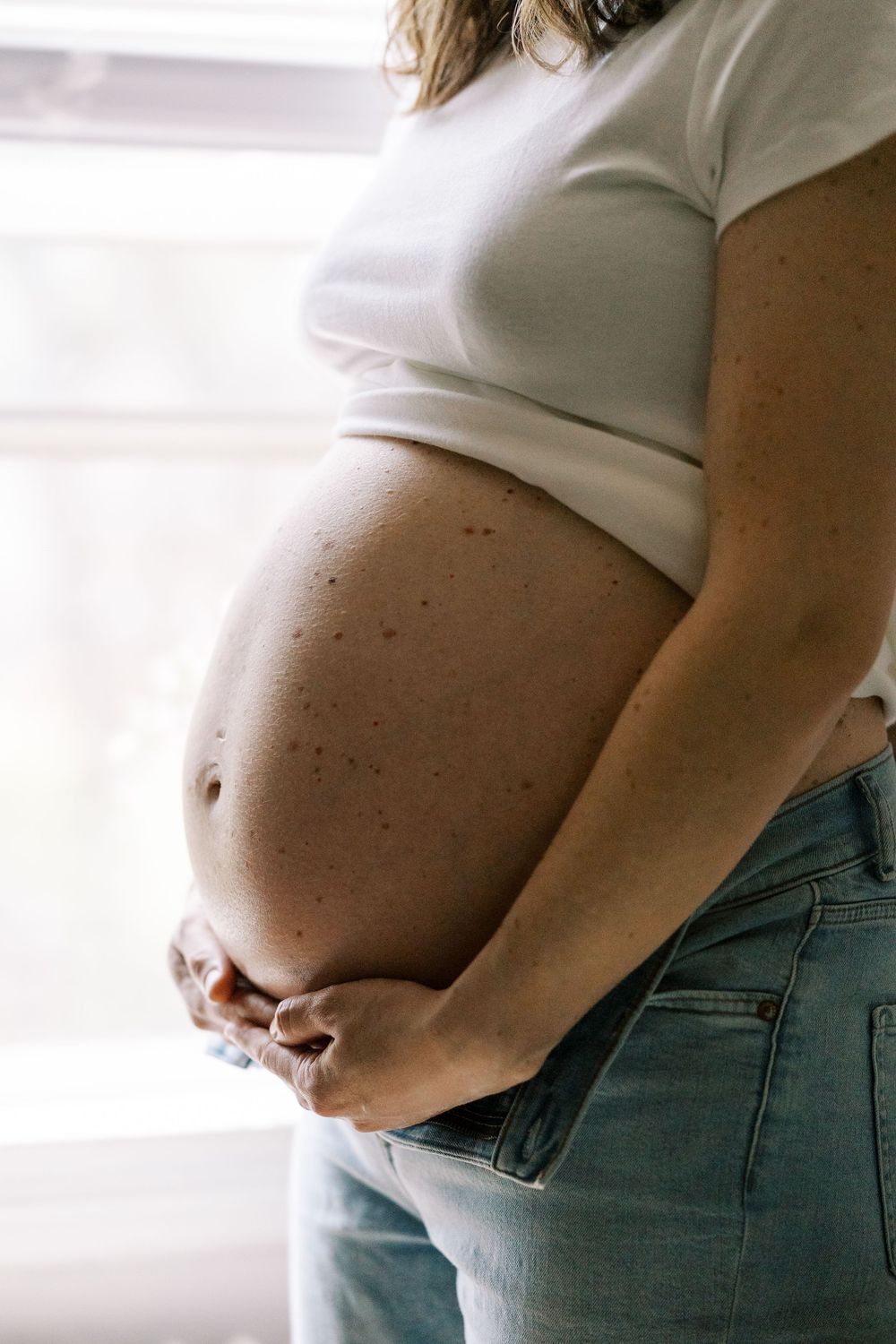 Side view of a pregnant belly in a white shirt and jeans against a bright window in Clifton, New Jersey.