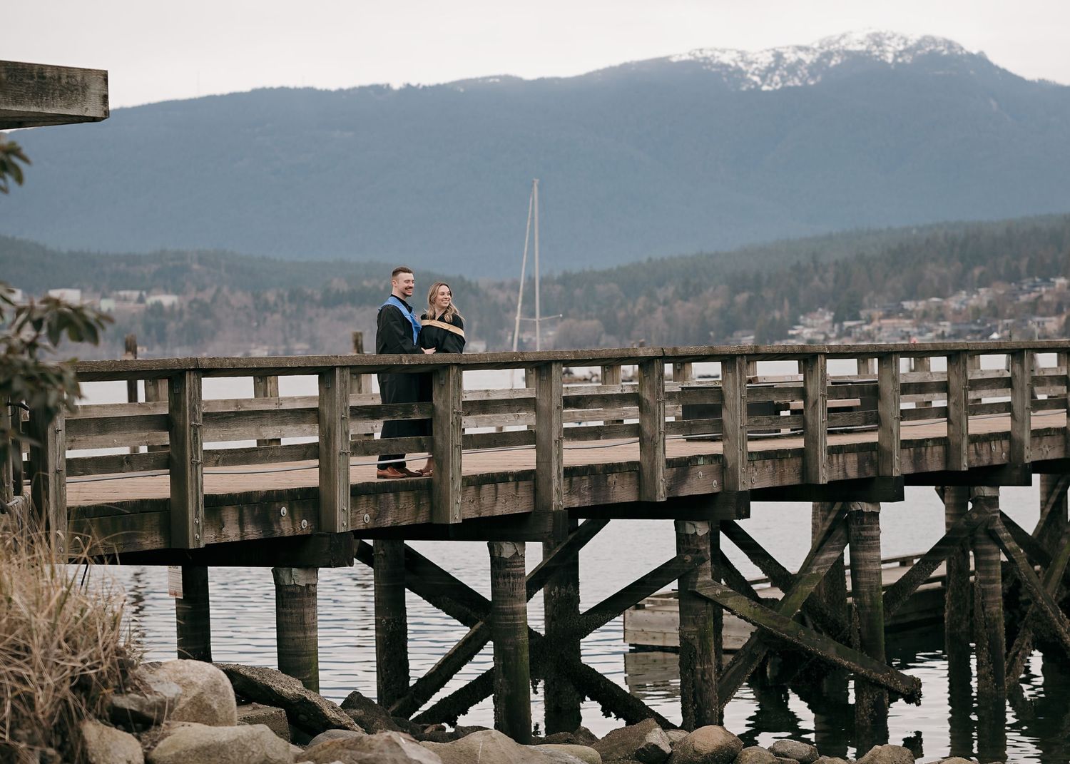 Someone sits on a wooden pier overlooking a scenic mountain landscape with calm waters and snow-capped peaks.