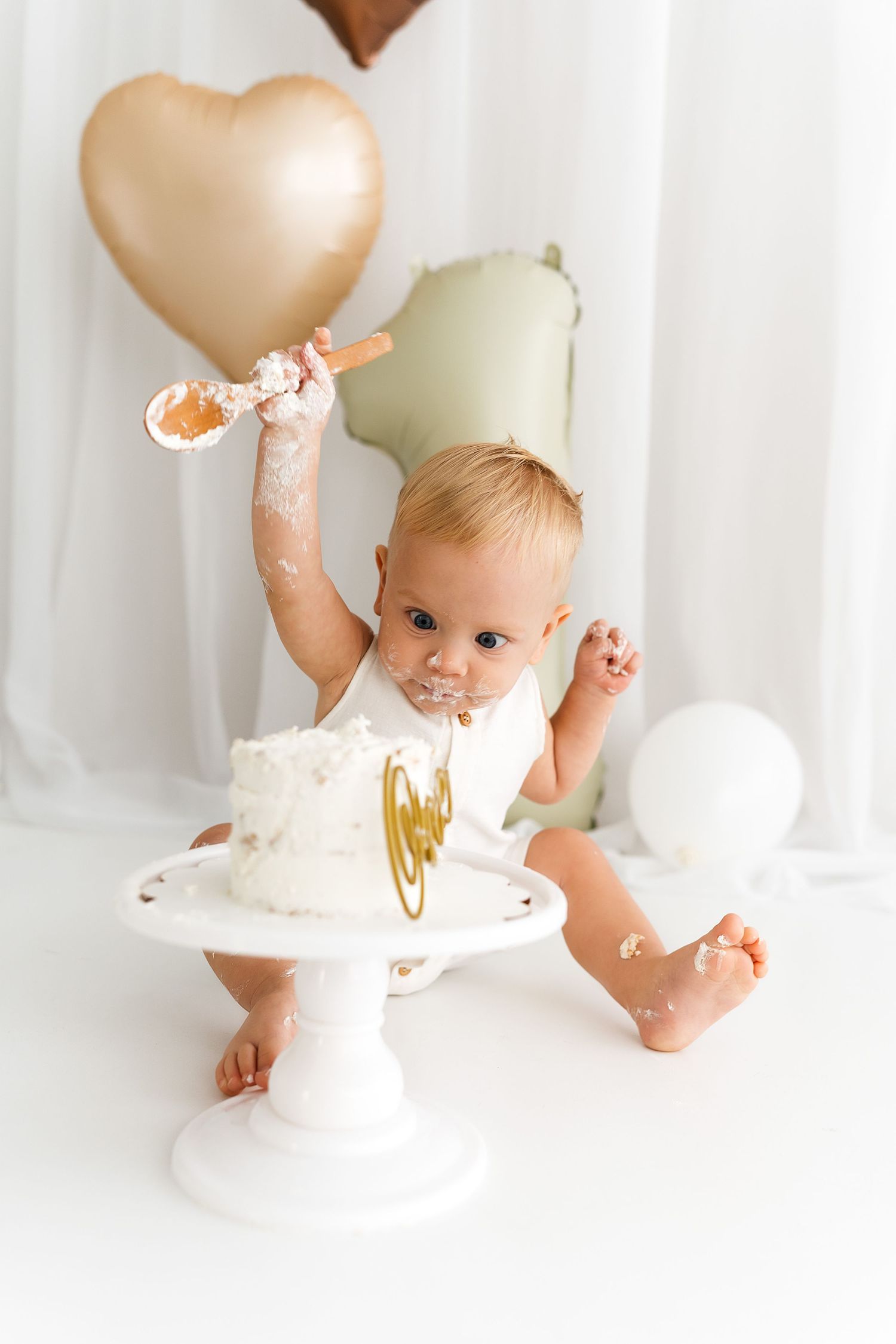 A baby sits beside a white cake stand during a first birthday celebration with heart-shaped balloons in the background.