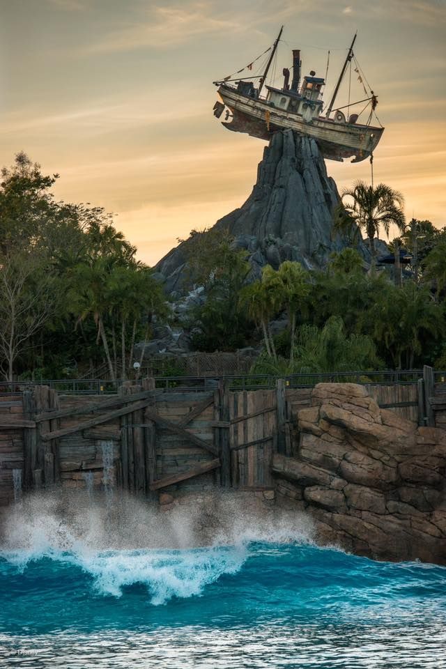 A shipwreck-themed water attraction creates dramatic waves crashing against rocky cliffs at sunset in a theme park.