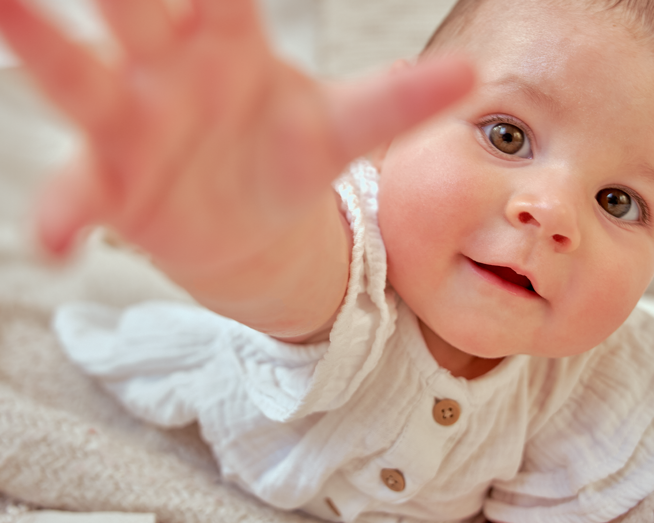 Baby in white dress reaching toward camera with bright expression.