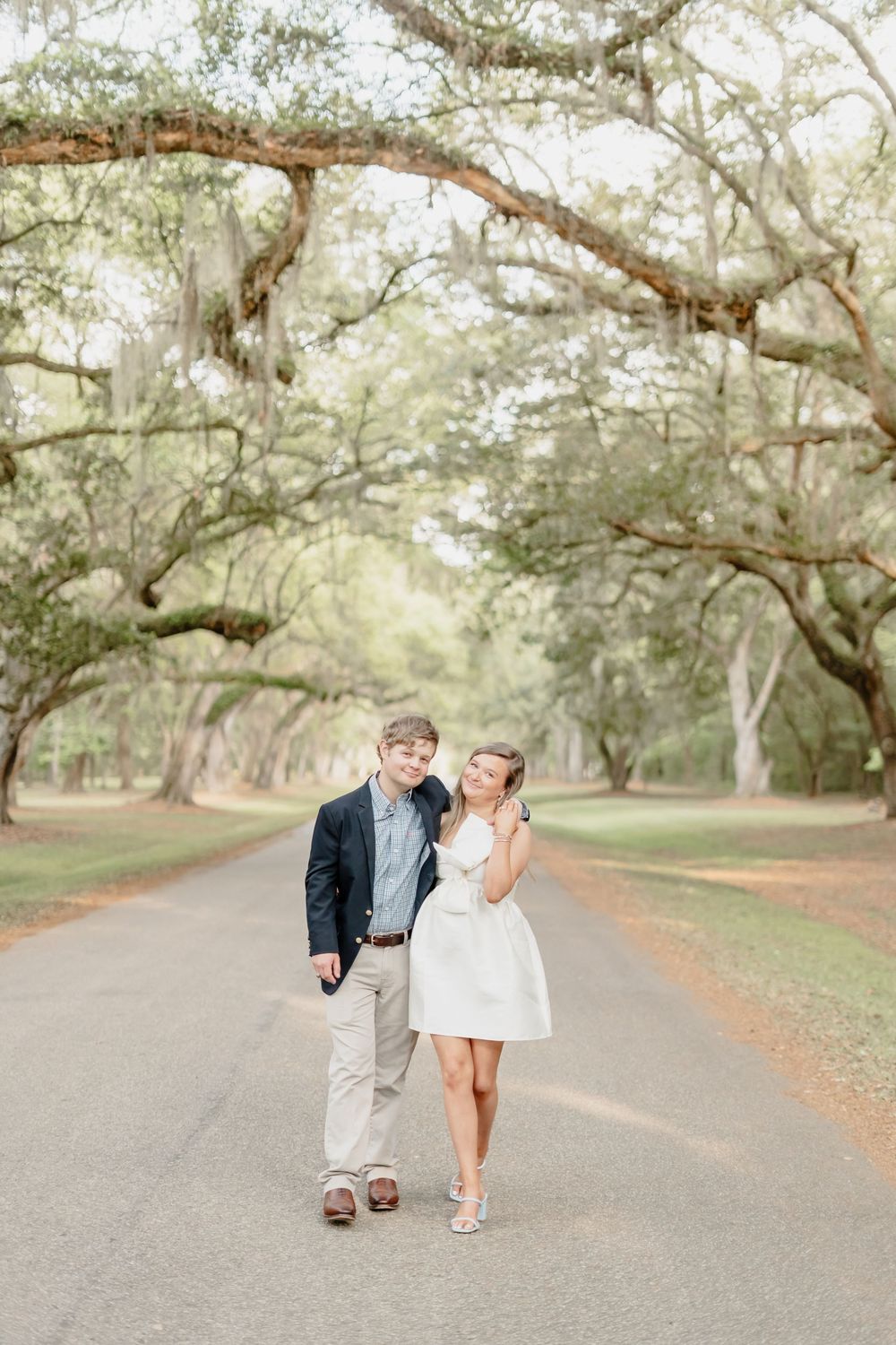 A couple walks down a dreamy oak-lined path together in coordinated dressy casual outfits.