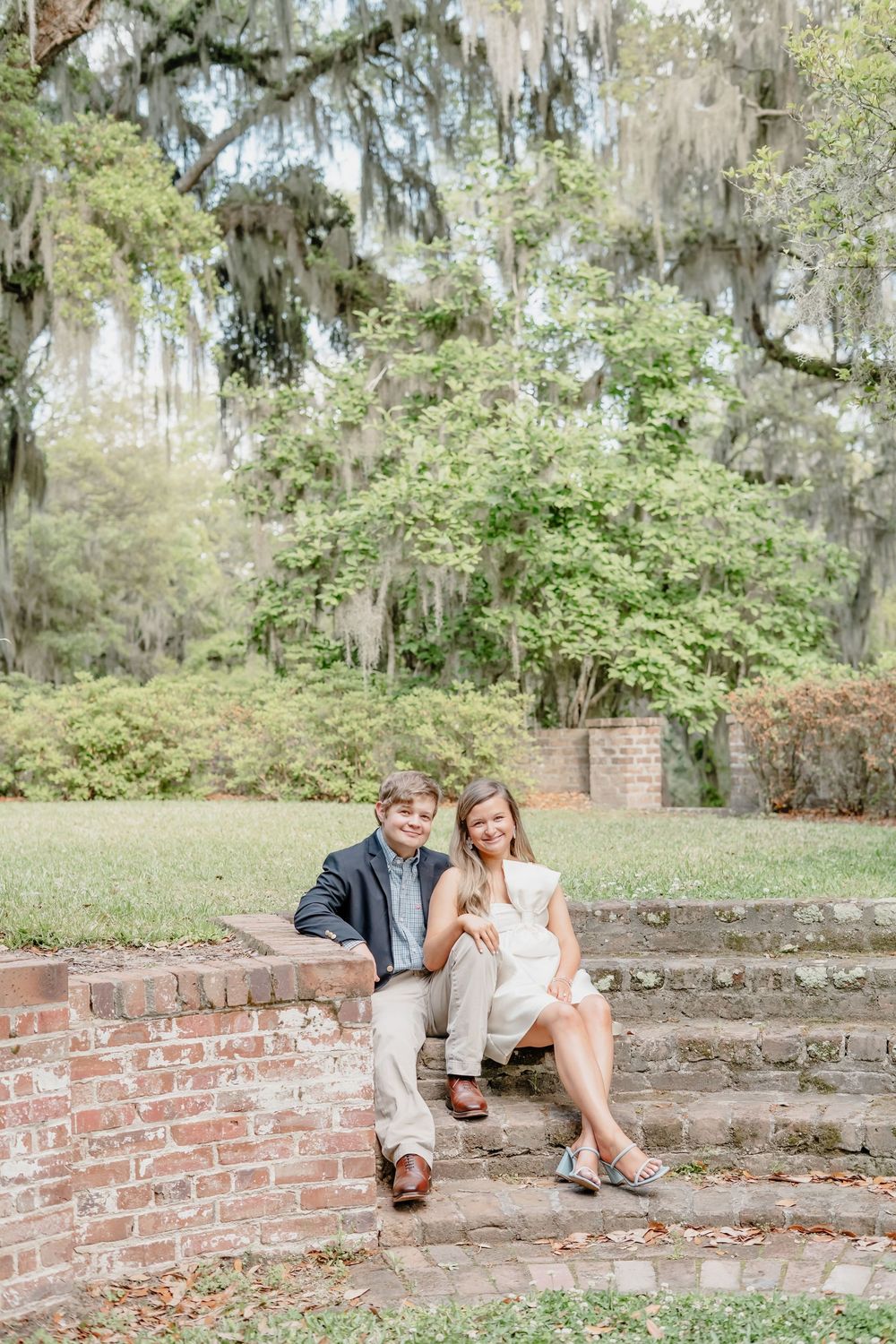 A romantic couple sits on brick stairs surrounded by Spanish moss and lush greenery in a Southern garden setting.