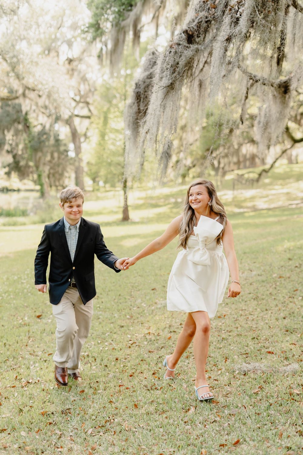 A couple holds hands while skipping through a garden beneath towering Spanish moss trees.