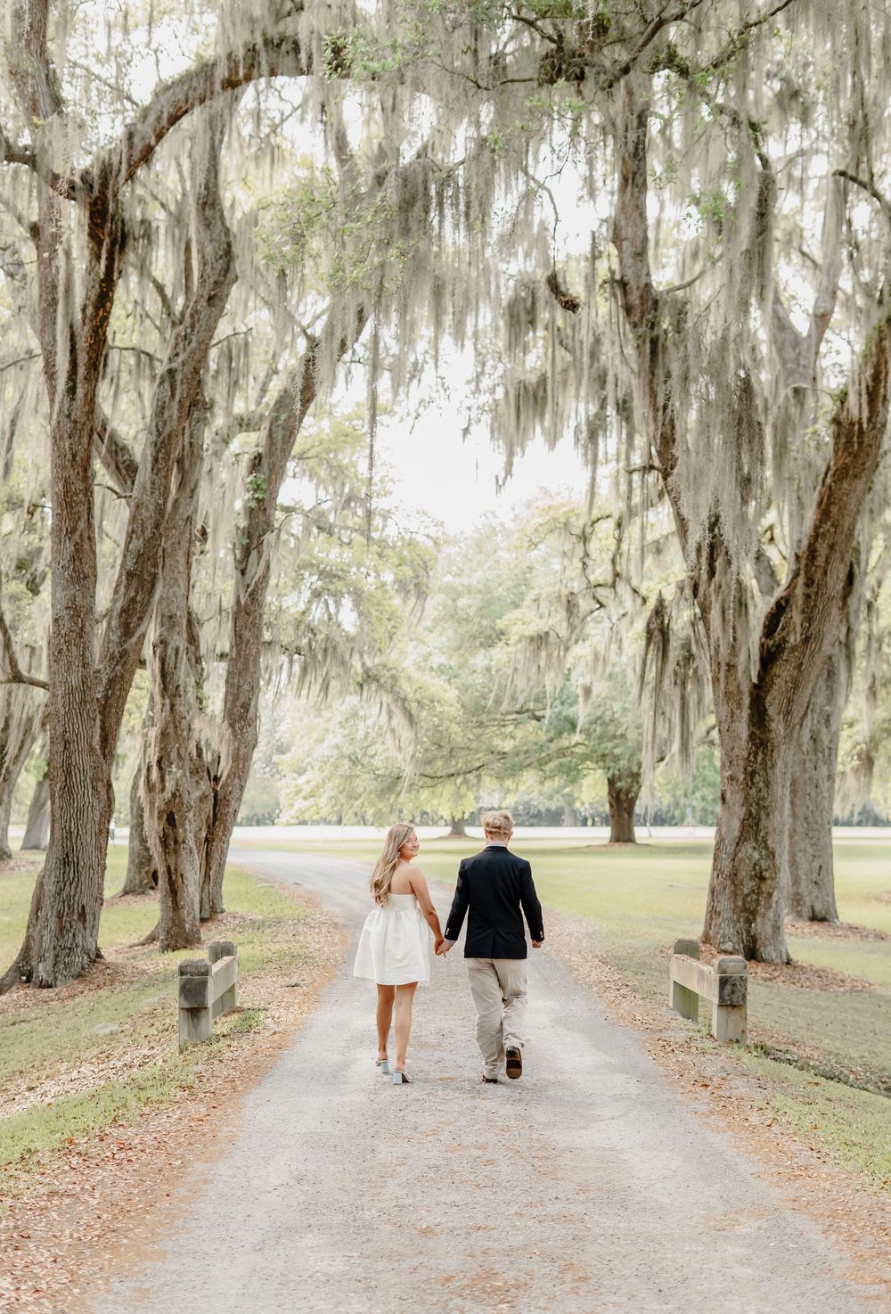 A couple walks hand in hand down a tree-lined path with hanging Spanish moss in a Southern garden.