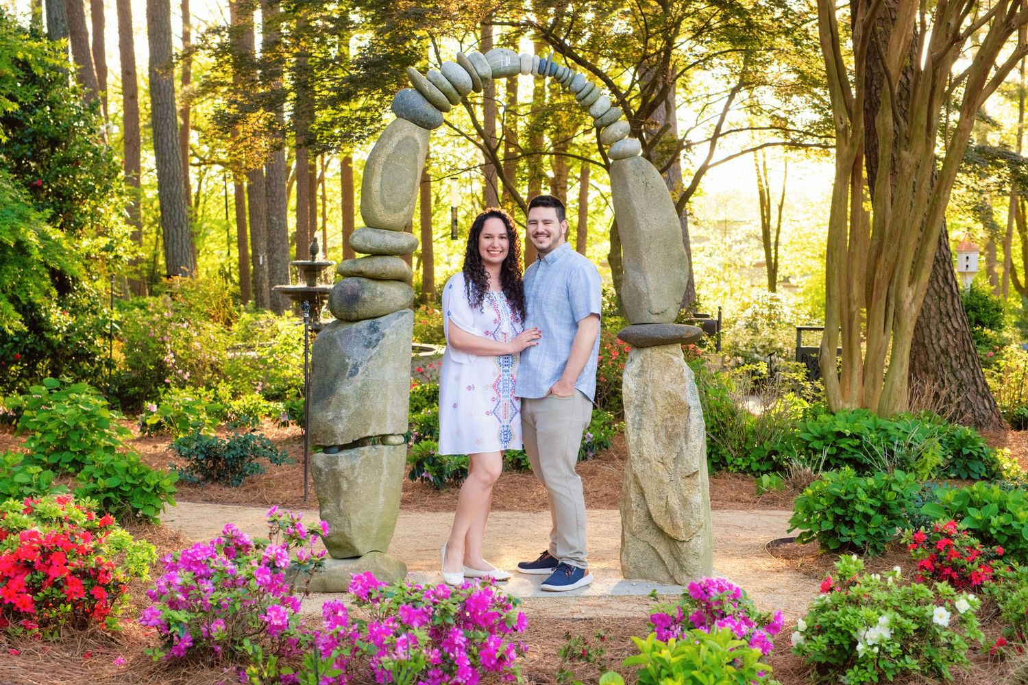 Couple stands beneath stone archway surrounded by colorful pink flowers and trees in park at sunset.