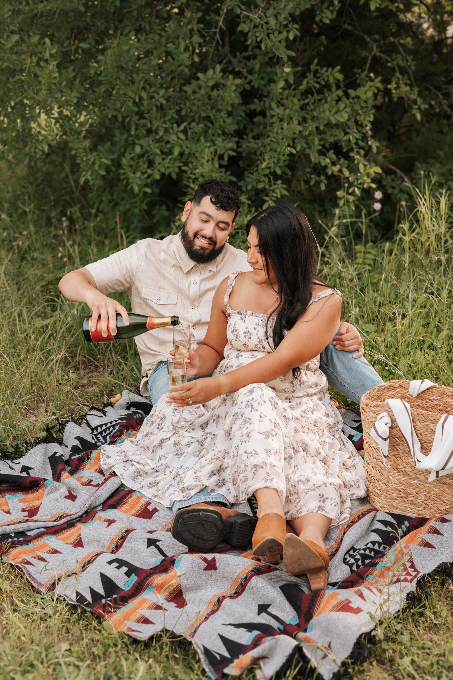 A couple enjoys a romantic picnic on a patterned blanket in a grassy outdoor setting with wine and snacks.