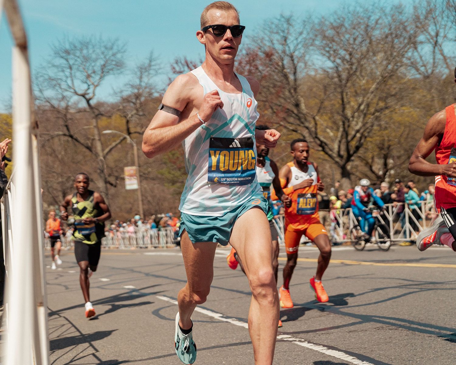 Marathon runner in light blue singlet and sunglasses competes in race through city park on sunny day.