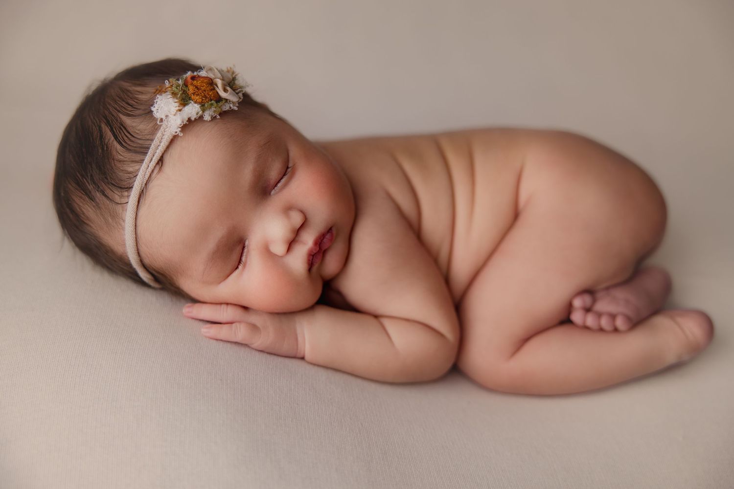 Peaceful newborn baby girl sleeping curled up, wearing a delicate floral headband, on a soft neutral backdrop.