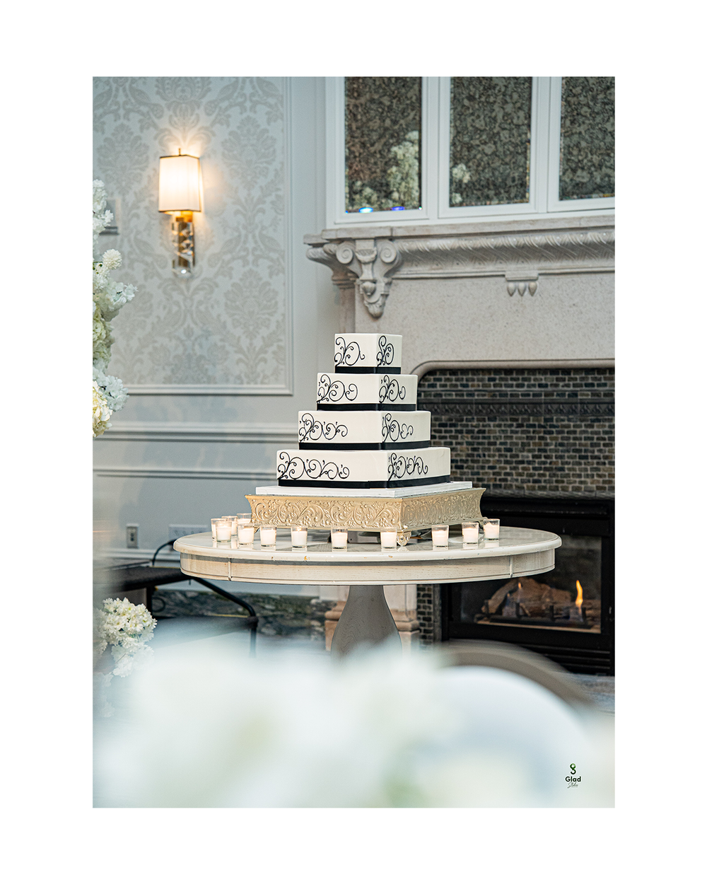 Elegant three-tier white wedding cake with black scroll details displayed in front of a modern fireplace.