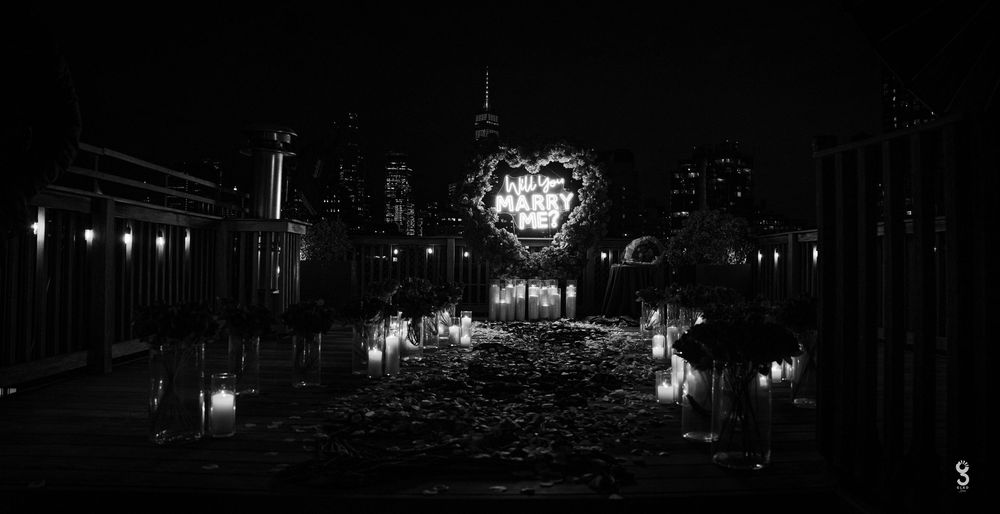 A black and white panoramic view of a dark street with illuminated buildings and trees at night.