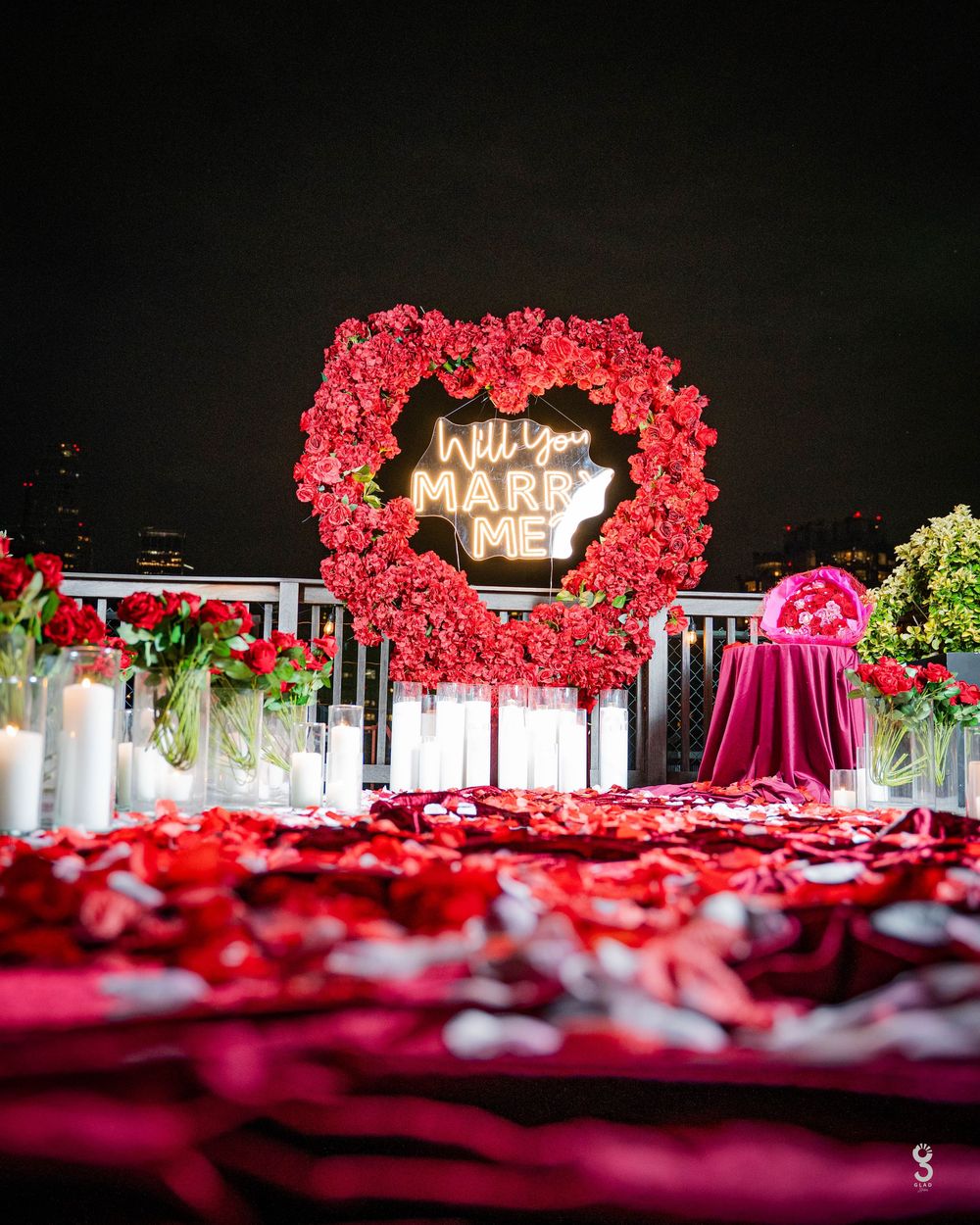 Illuminated 'MARRY' sign glows above a romantic red rose heart-shaped arch against a dark nighttime backdrop.