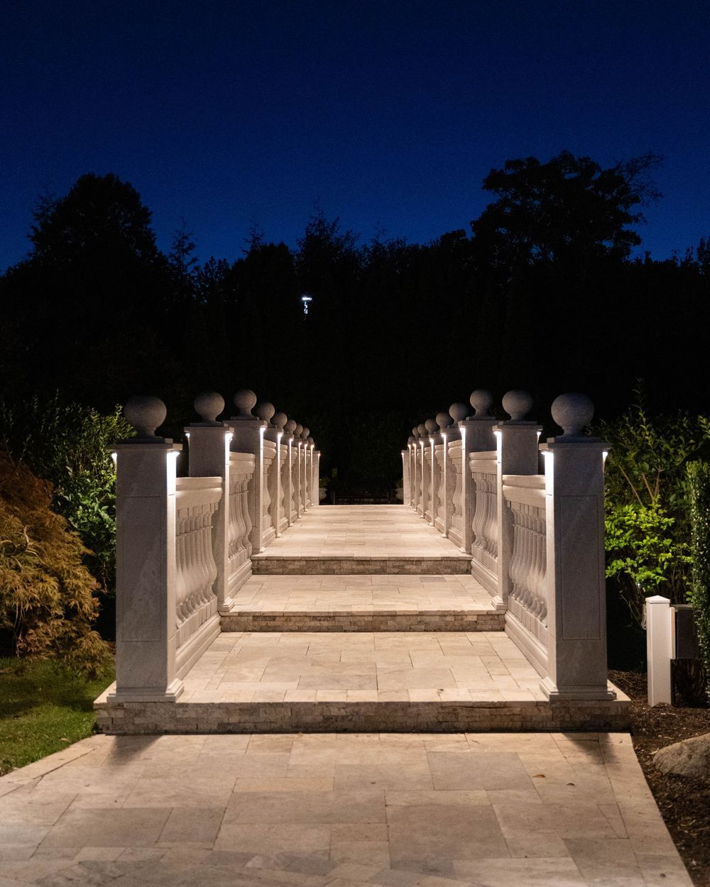 A symmetrical concrete walkway with white pillars and lighting fixtures extends into darkness at night.