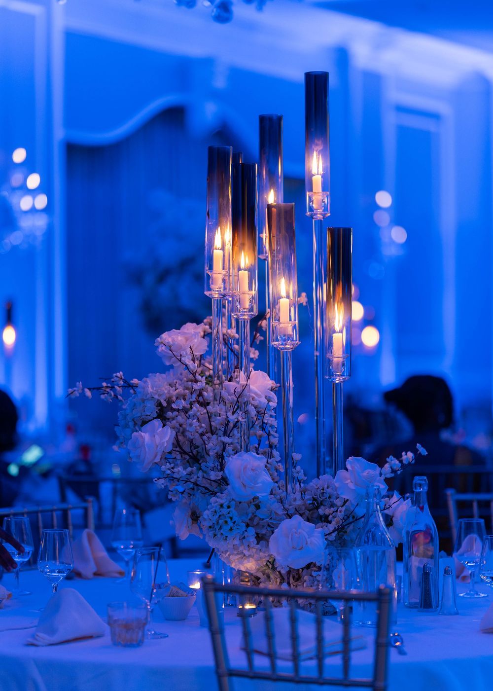 Elegant wedding reception tables decorated with tall glass candelabras and white floral centerpieces under blue lighting.