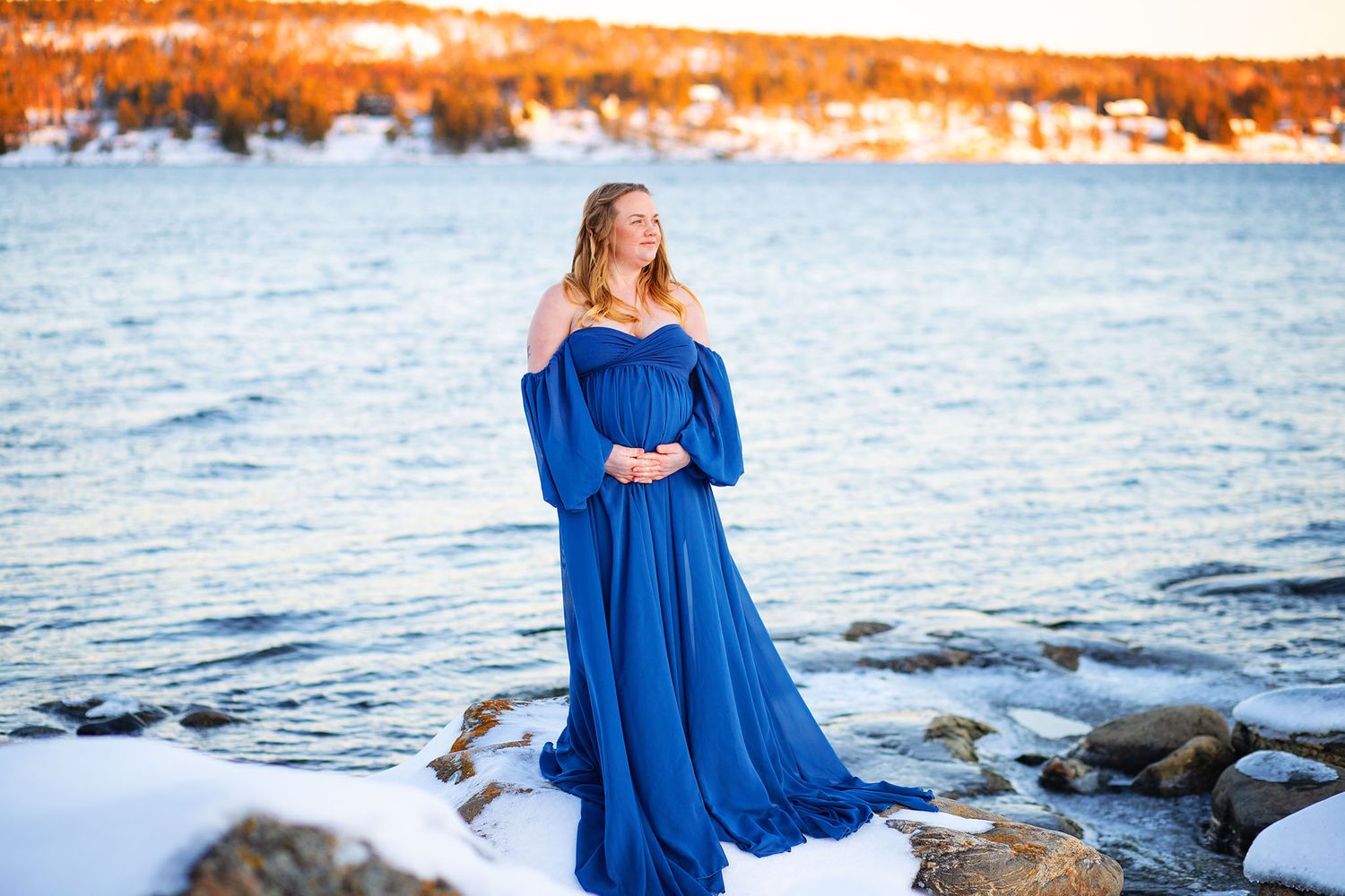 Pregnant woman in royal blue off-shoulder gown on snowy rocky winter shoreline.