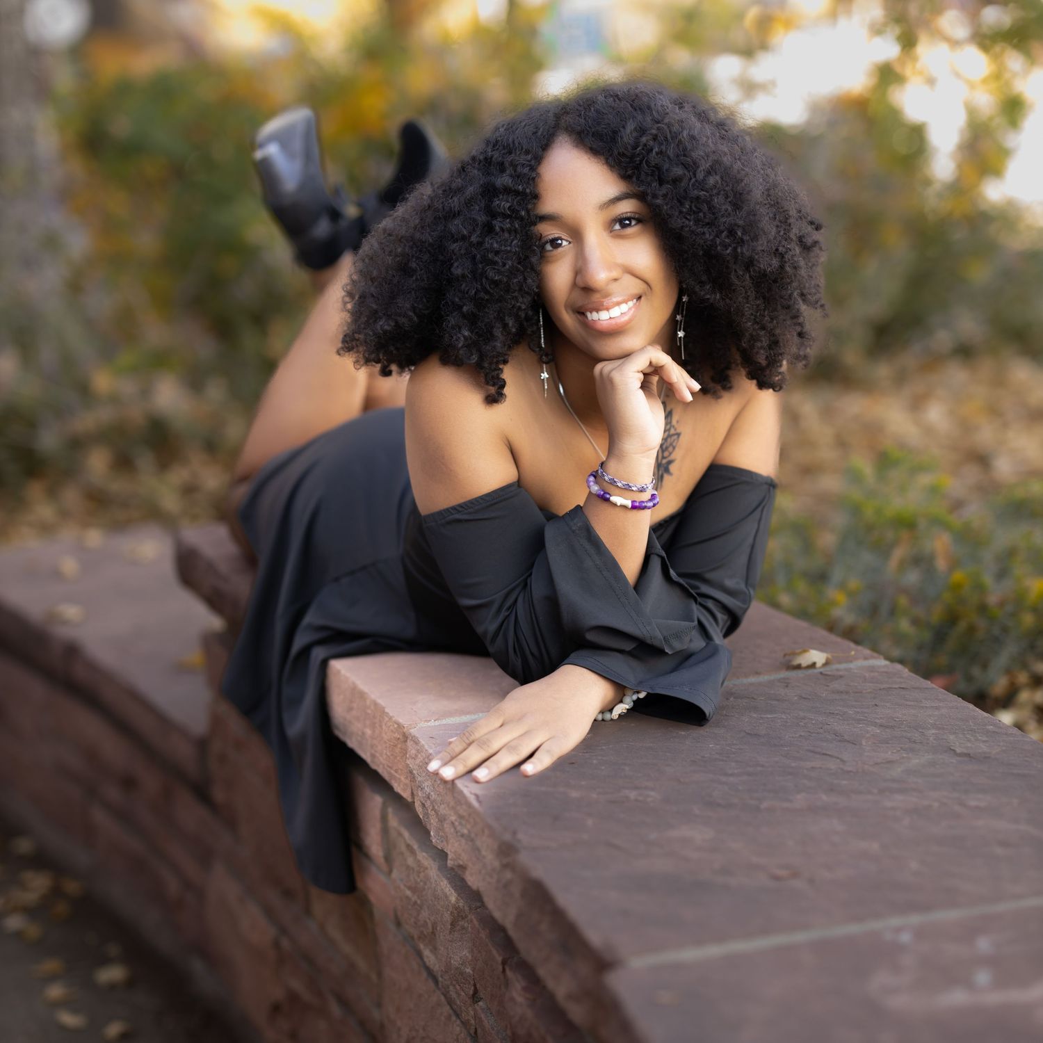 A person in a black off-shoulder top lounges on a wooden surface outdoors, showcasing natural curly hair and jewelry.