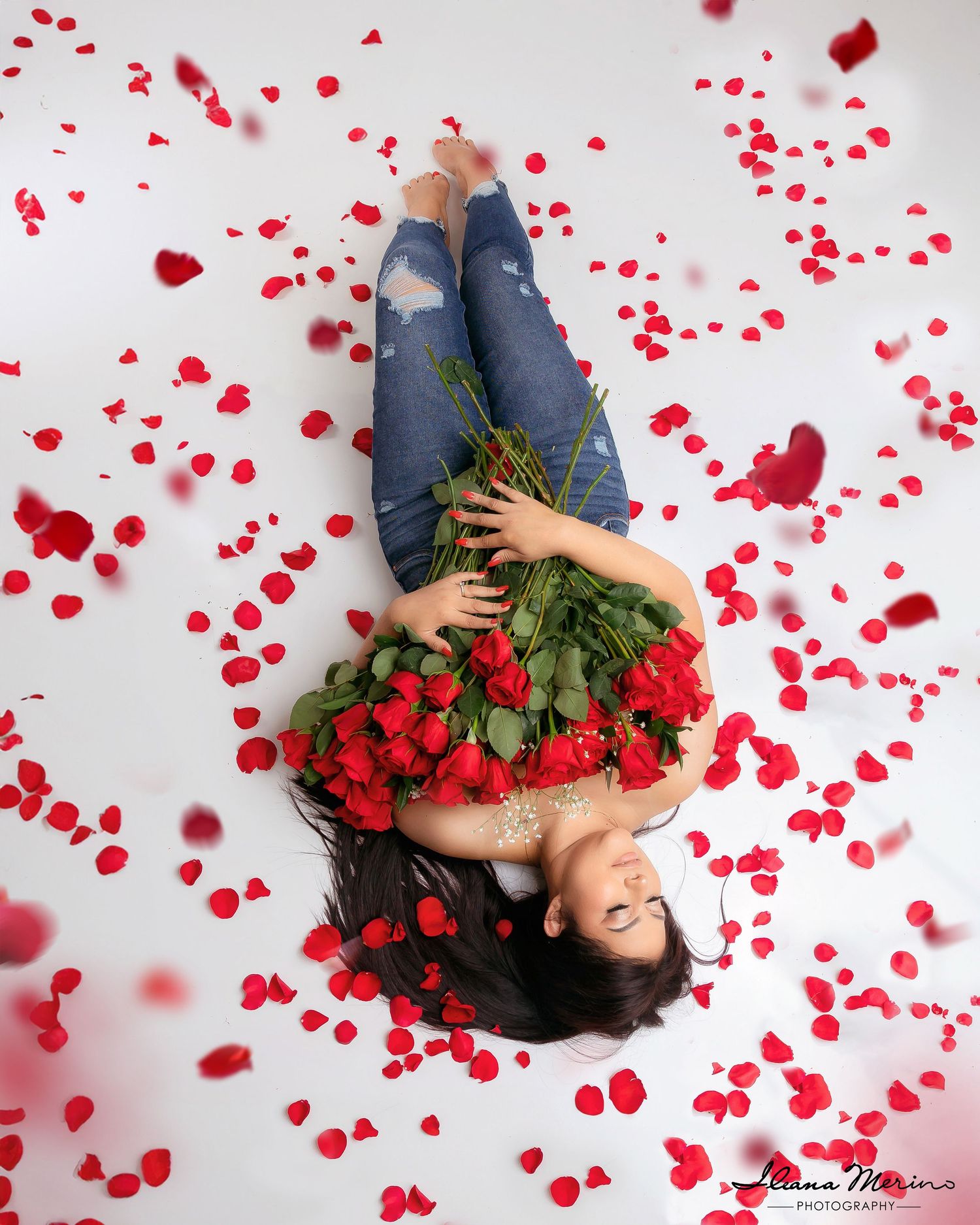 Woman lying on white floor surrounded by red rose petals holding a bouquet of red roses from above.