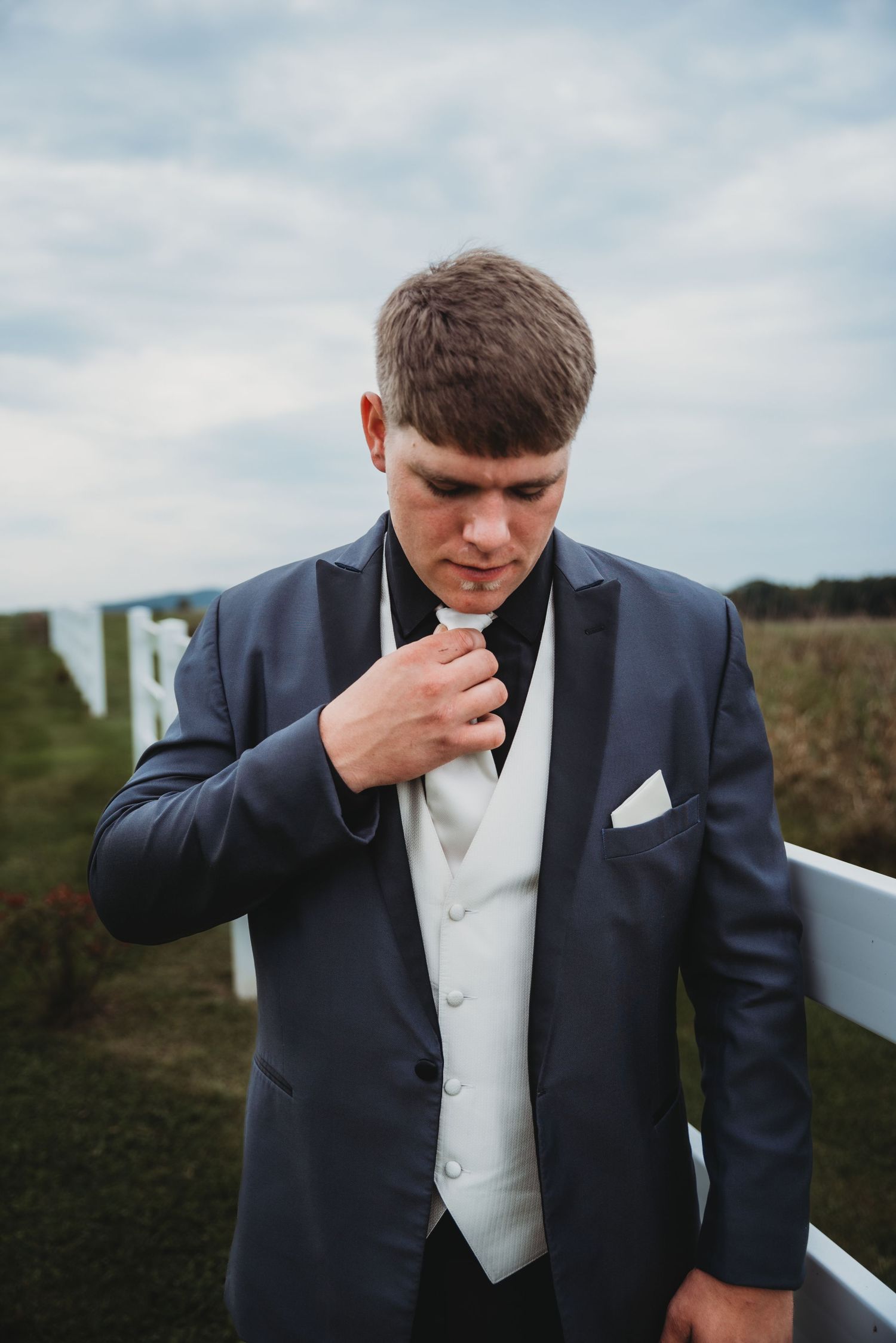 Groom in navy suit and white tie adjusts his collar outdoors.
