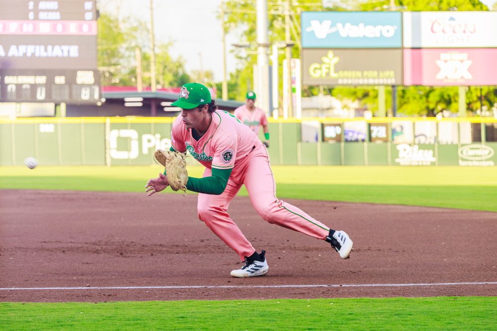 Baseball player in pink uniform and green cap fielding a ground ball on a dirt infield during a game.