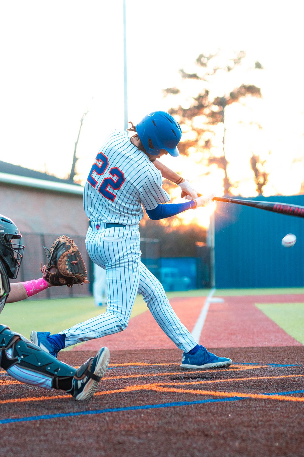 Baseball player in striped uniform swings bat at sunset during game on artificial turf field.