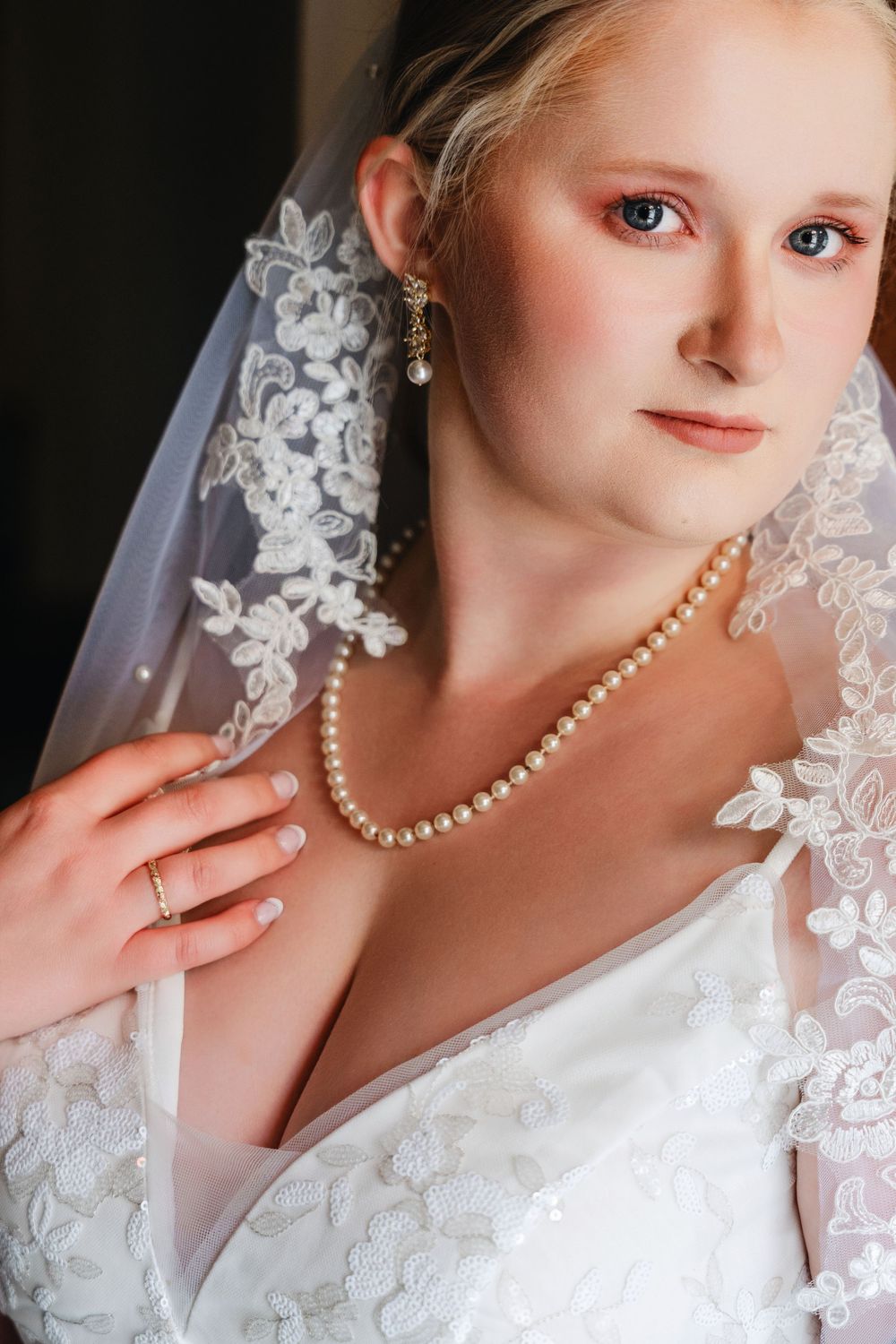 A bride wearing a white lace wedding dress with delicate pearl necklace and floral veil details.