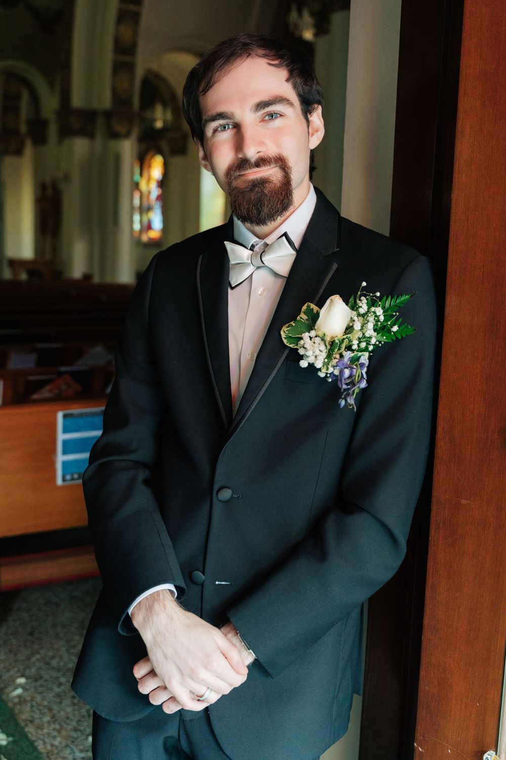 A groom in a black tuxedo and bow tie stands in a church with a floral boutonniere featuring white calla lily and greenery.