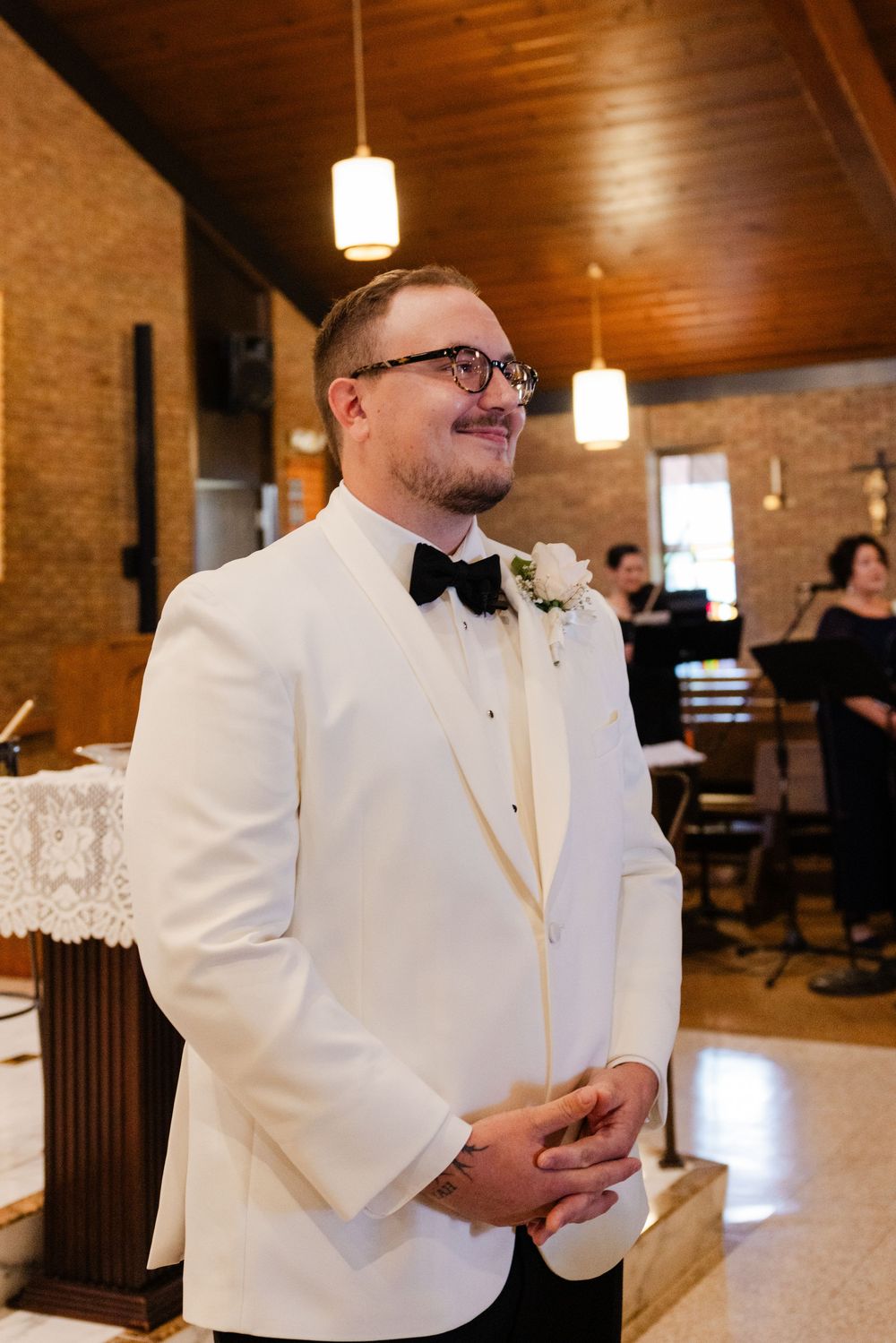 A groom in a white tuxedo jacket, black bow tie and glasses smiles while standing at the altar in a brick-walled church.