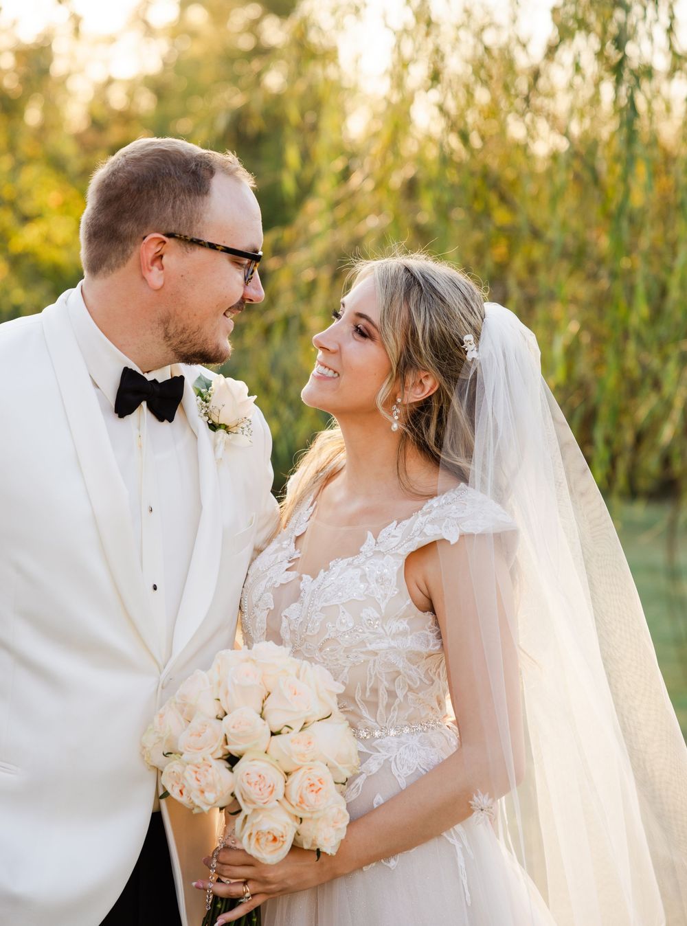 A newlywed couple shares an intimate moment outdoors with soft sunset lighting and a bouquet of blush roses.