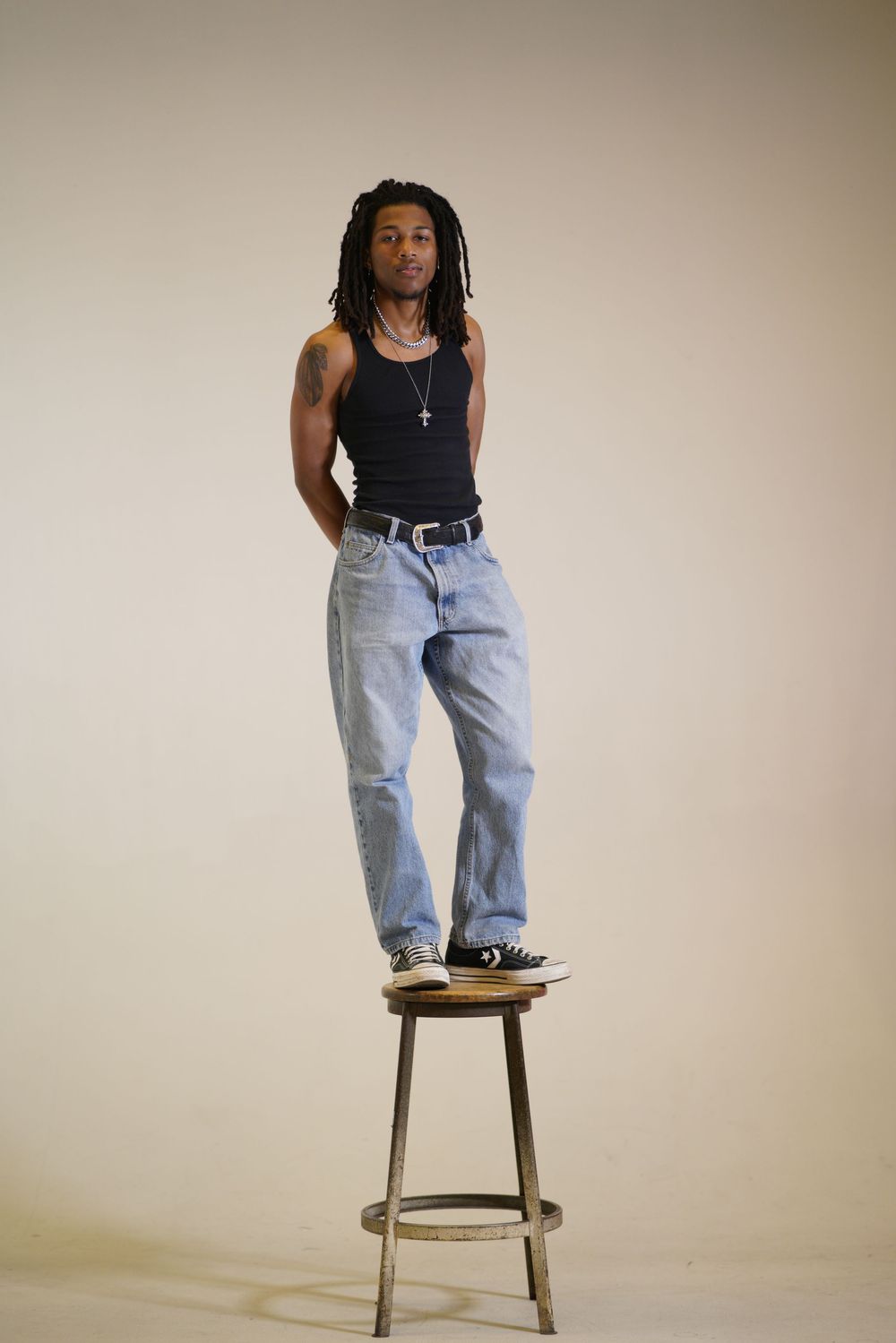 Person in black tank top and light wash jeans stands confidently on metal stool against neutral backdrop.