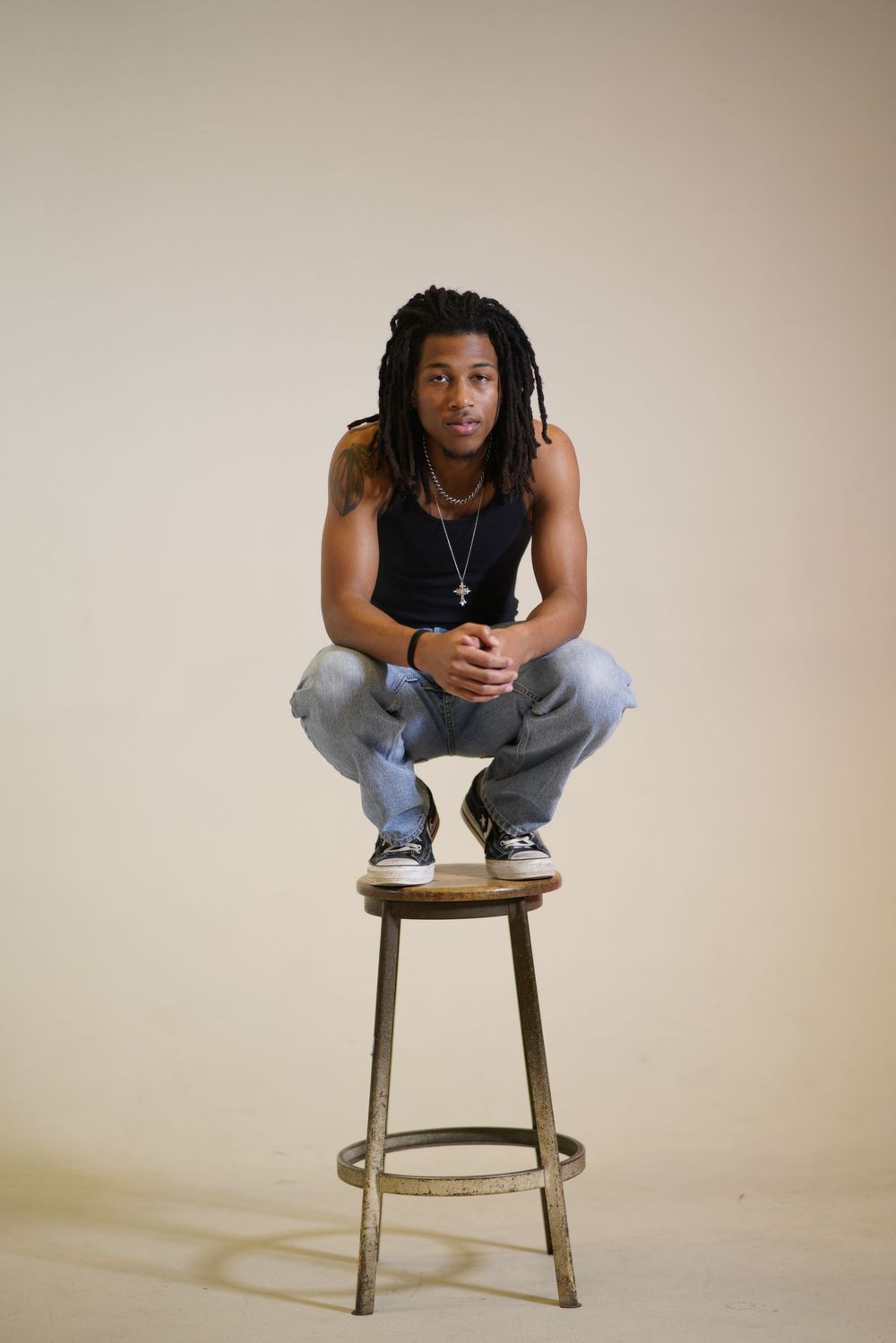 Person in black tank top and jeans crouches on metal barstool against neutral background in studio portrait.