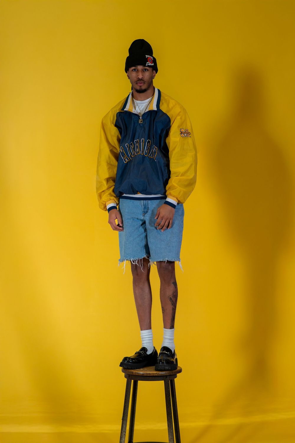 Person standing on black stool wearing yellow and blue varsity jacket with denim shorts against yellow background.