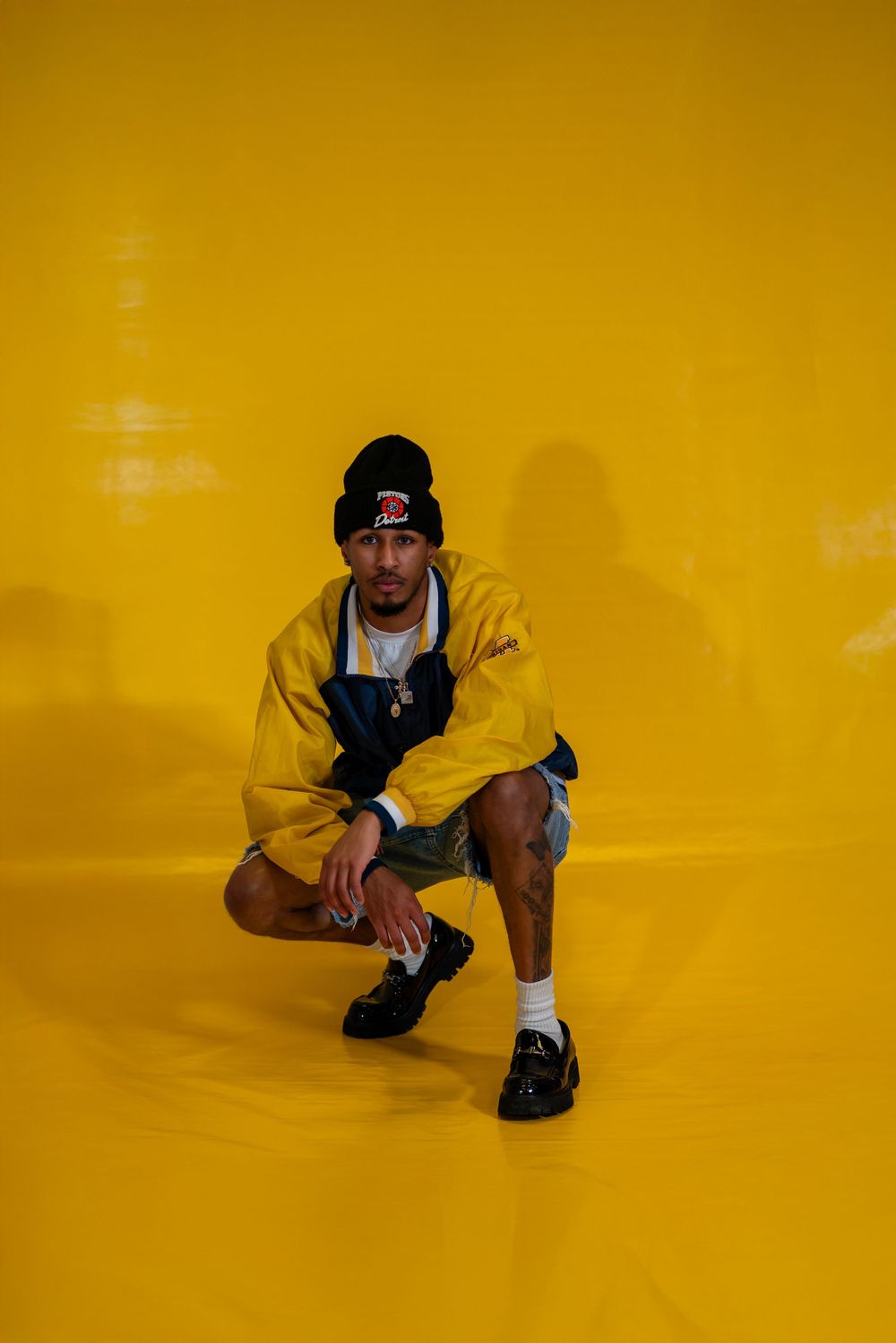 Fashion model in yellow hoodie and overalls crouches against vibrant yellow background in urban streetwear look.
