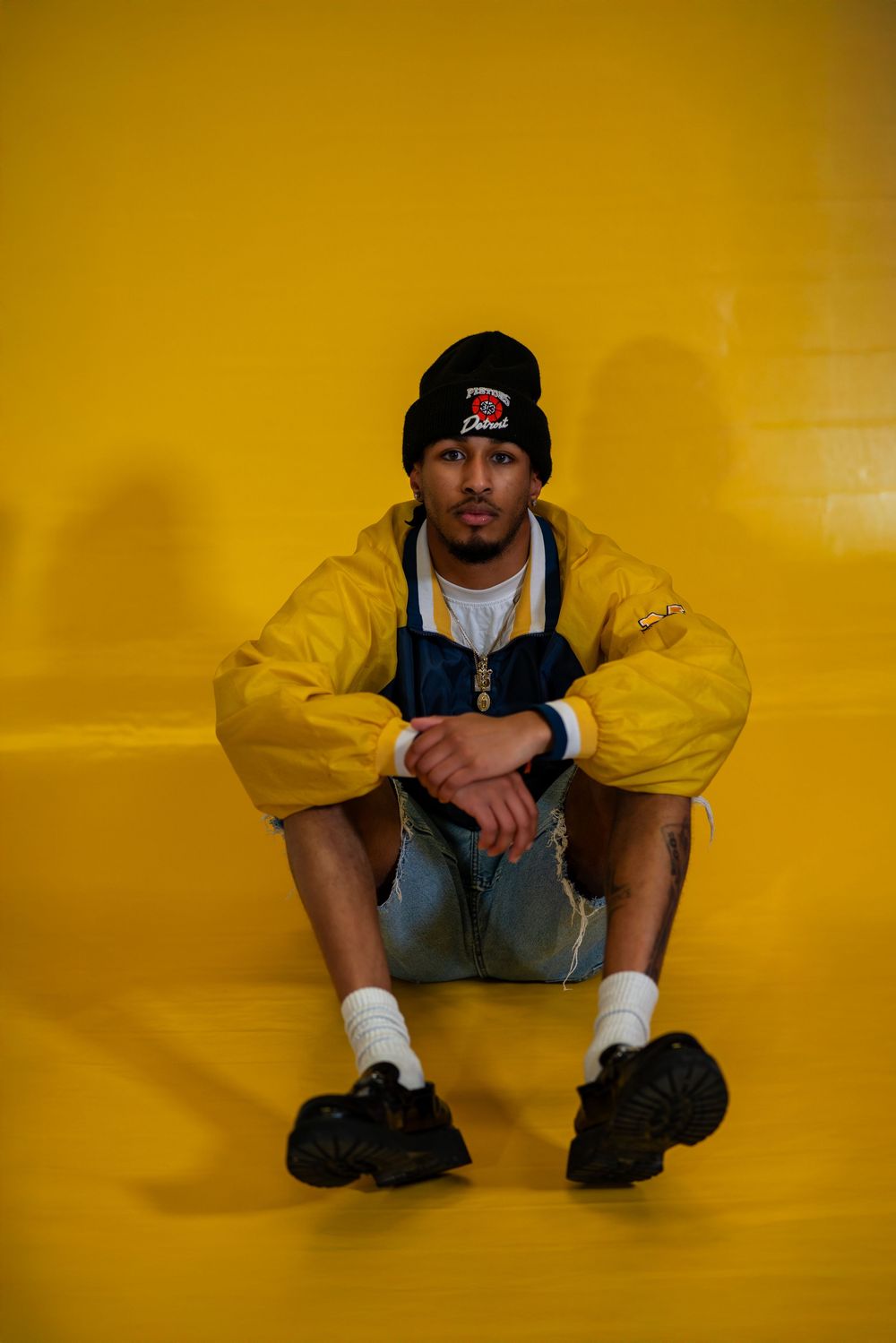 Person wearing yellow jacket and overalls sits against yellow wall while wearing a black beanie and white socks.