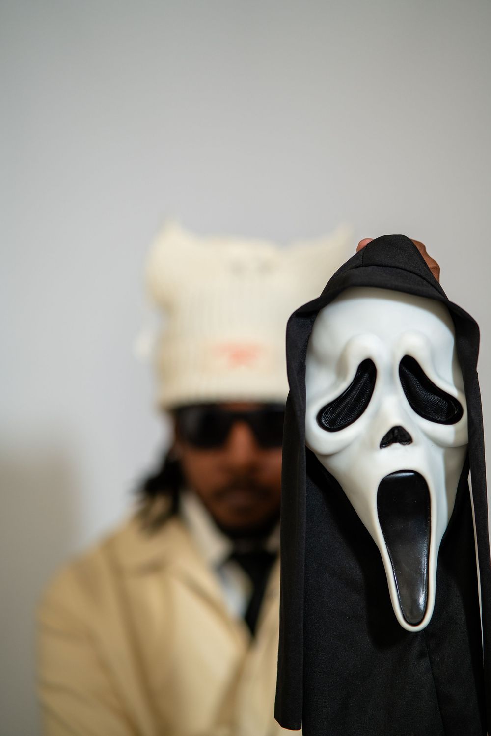A Ghostface mask from the Scream movie franchise stands in focus with a blurred figure wearing sunglasses in the background.