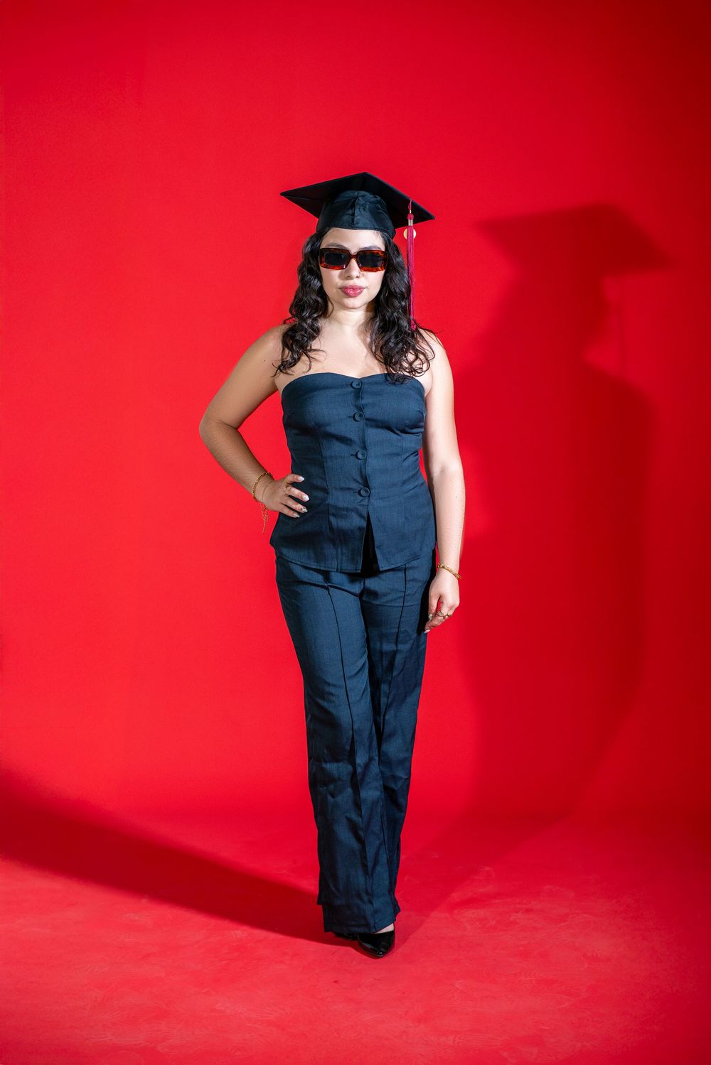 Graduate wearing navy blue jumpsuit and sunglasses poses against vibrant red background during commencement photo shoot.