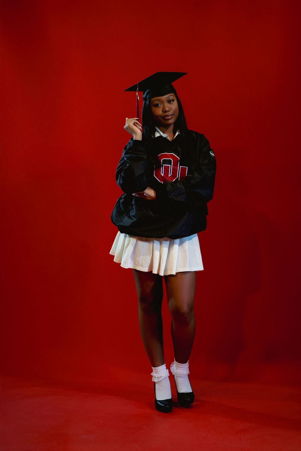 Graduate in black varsity jacket and cap poses against vibrant red backdrop with white pleated skirt and socks.