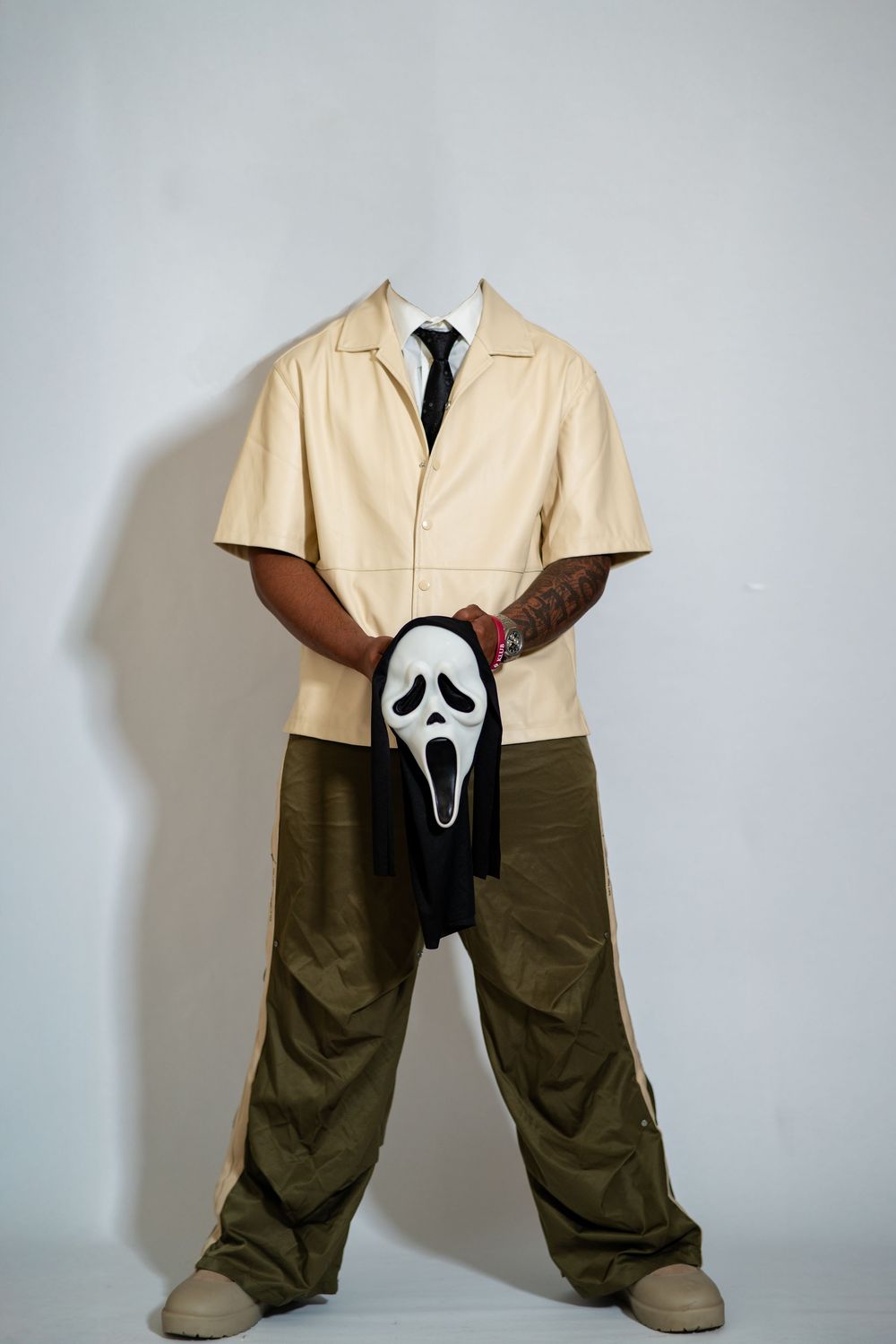 Figure wearing beige short-sleeve jacket and dark pants holds Ghostface mask from horror movie Scream.