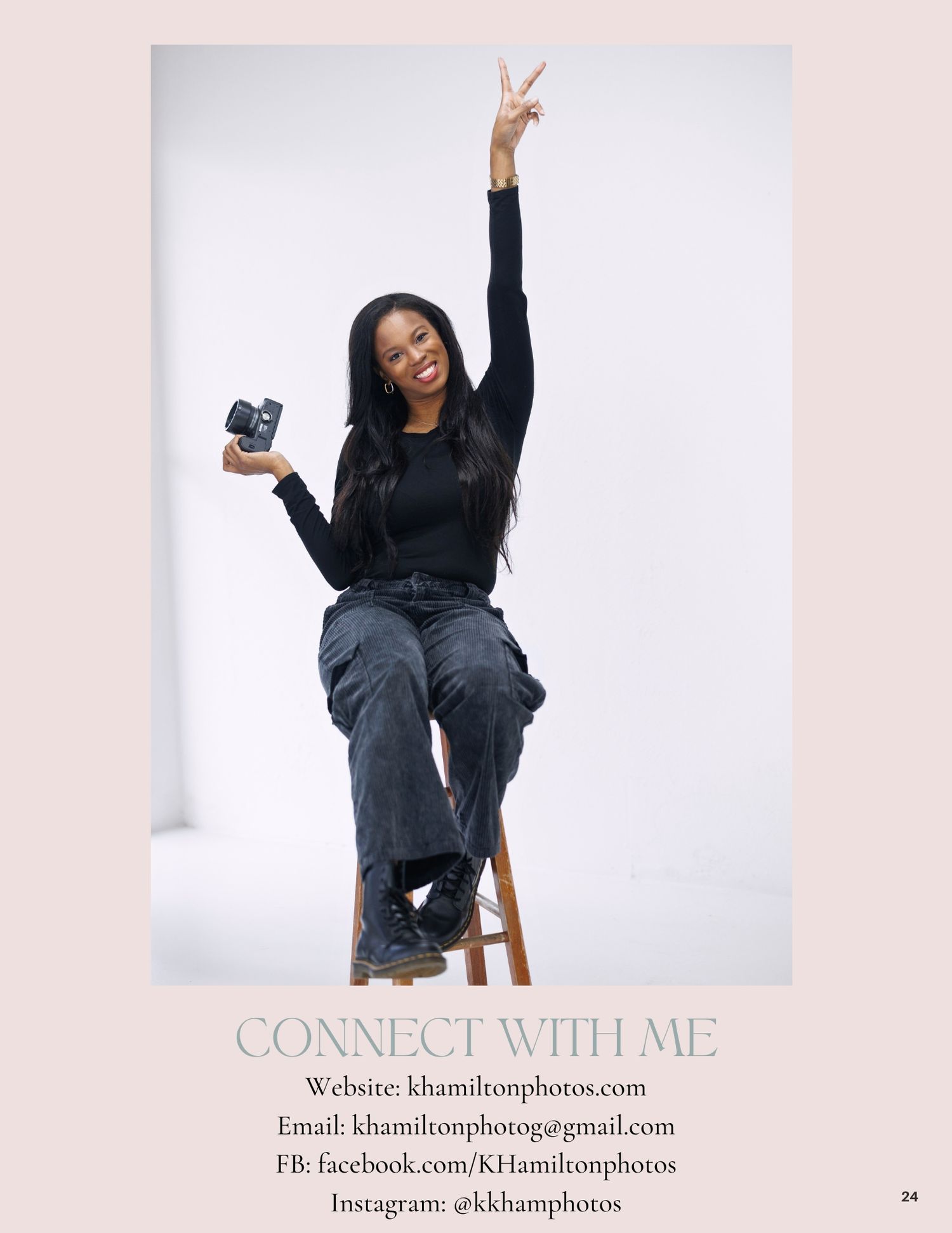 A photographer sits on a stool making peace sign while holding camera against white background.