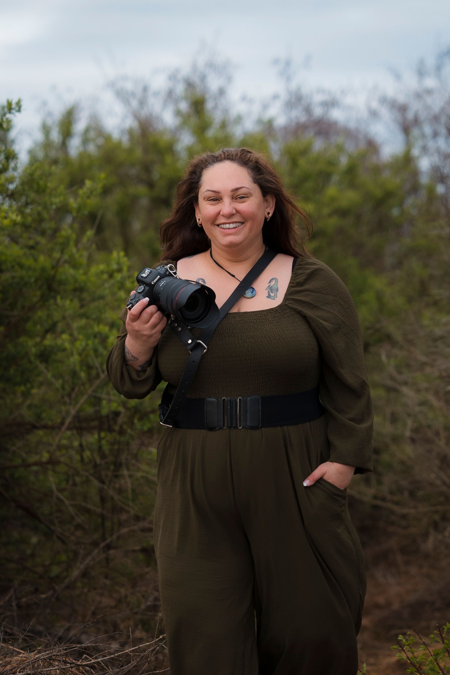 A photographer wearing an olive jumpsuit holds a camera while standing outdoors among green foliage.