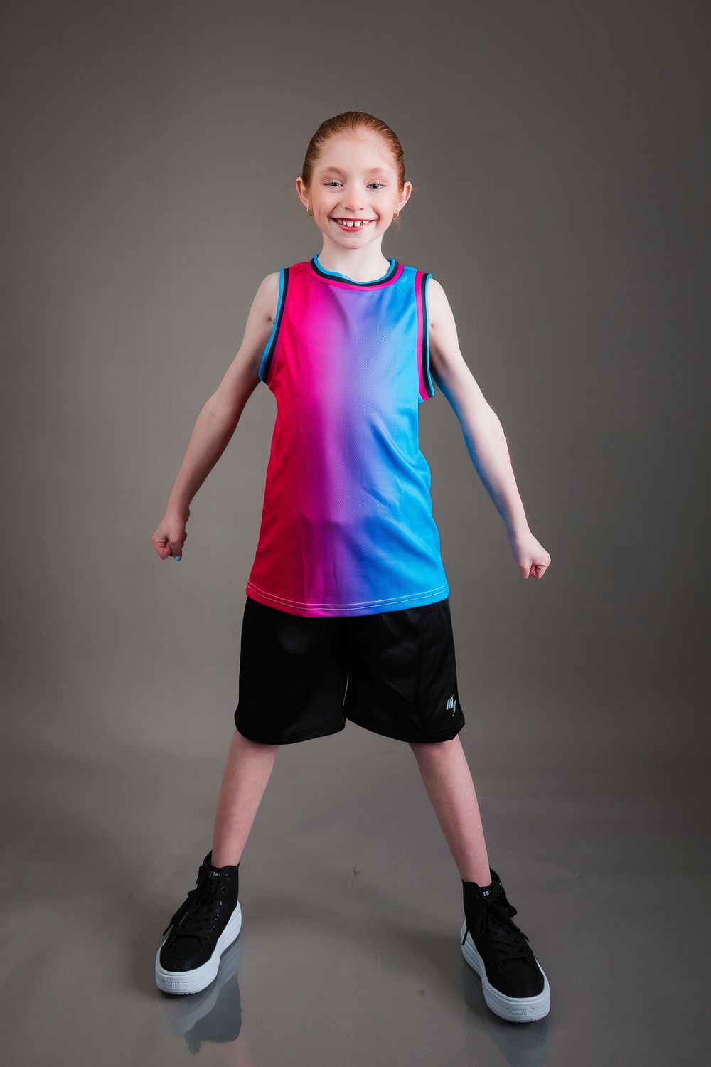 Young athlete in vibrant gradient tank top and black shorts poses energetically against gray studio background.