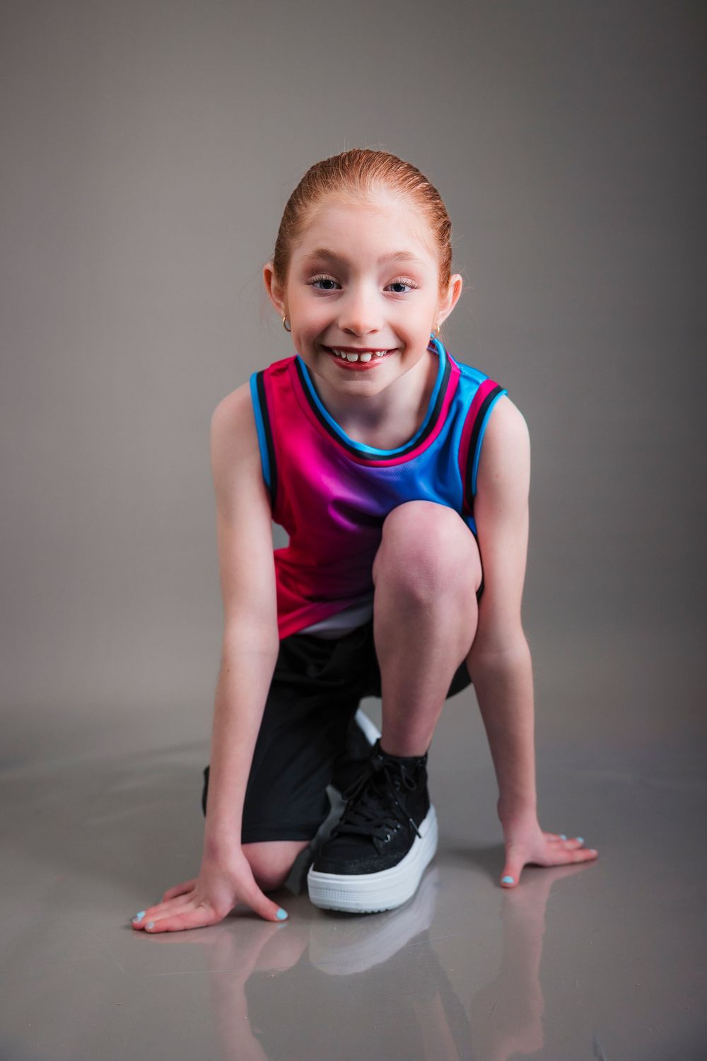Young athlete in pink and blue athletic wear crouches on a gray backdrop in a dynamic pose with a bright smile.