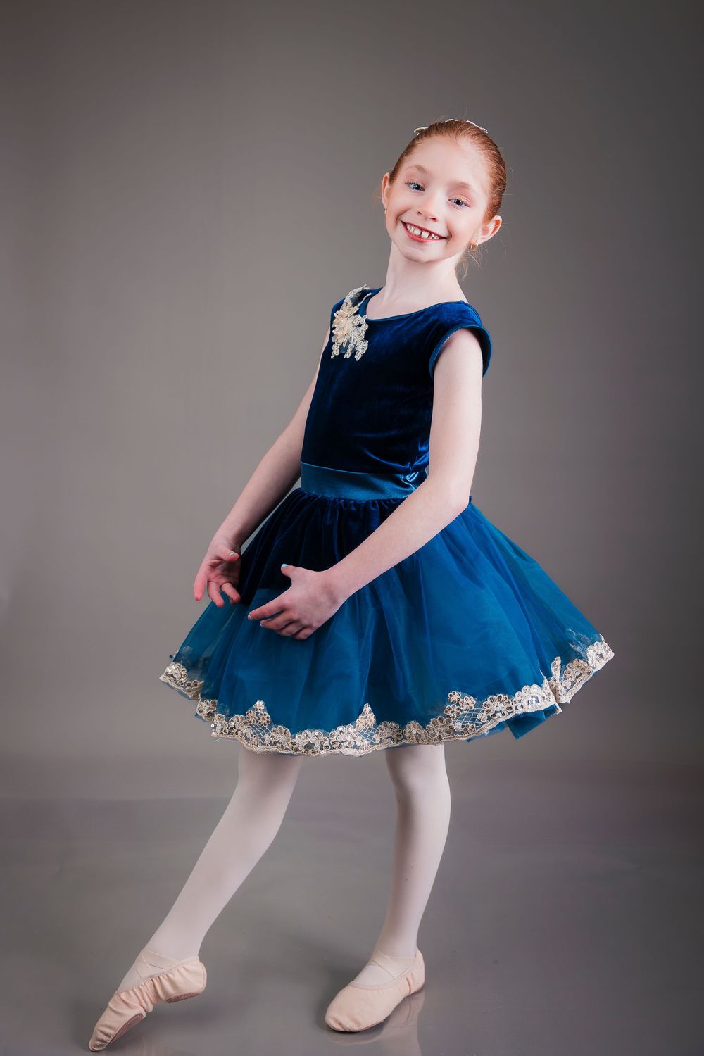 Young ballet dancer in blue velvet dress with tulle skirt and silver trim poses gracefully in dance position.