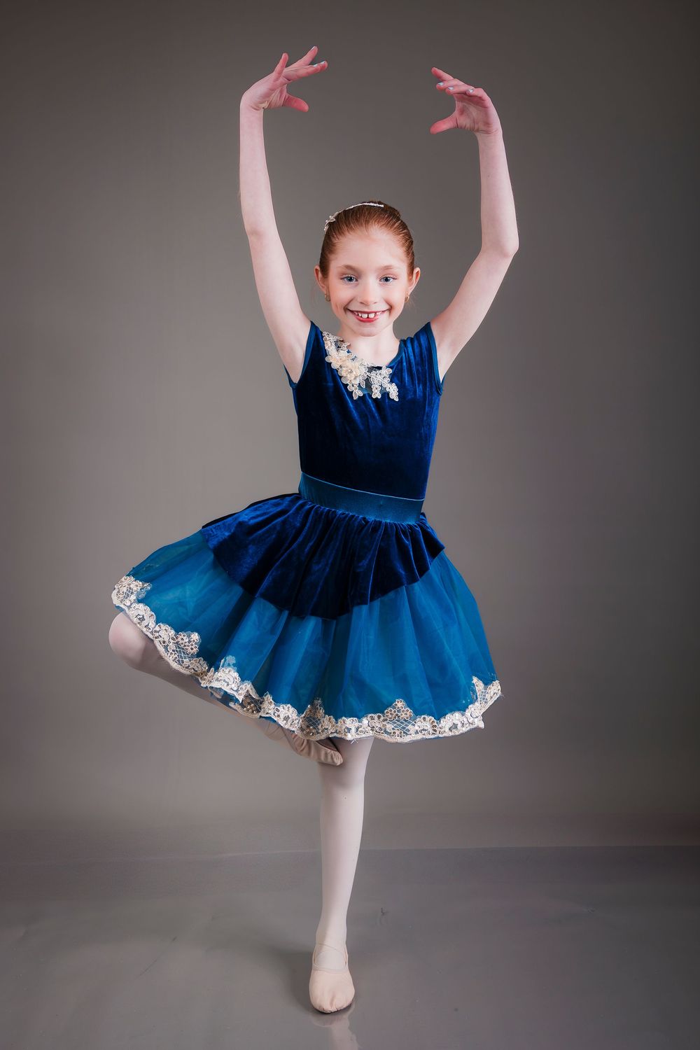 Young dancer in royal blue velvet tutu dress strikes a graceful ballet pose with raised arms against gray backdrop.