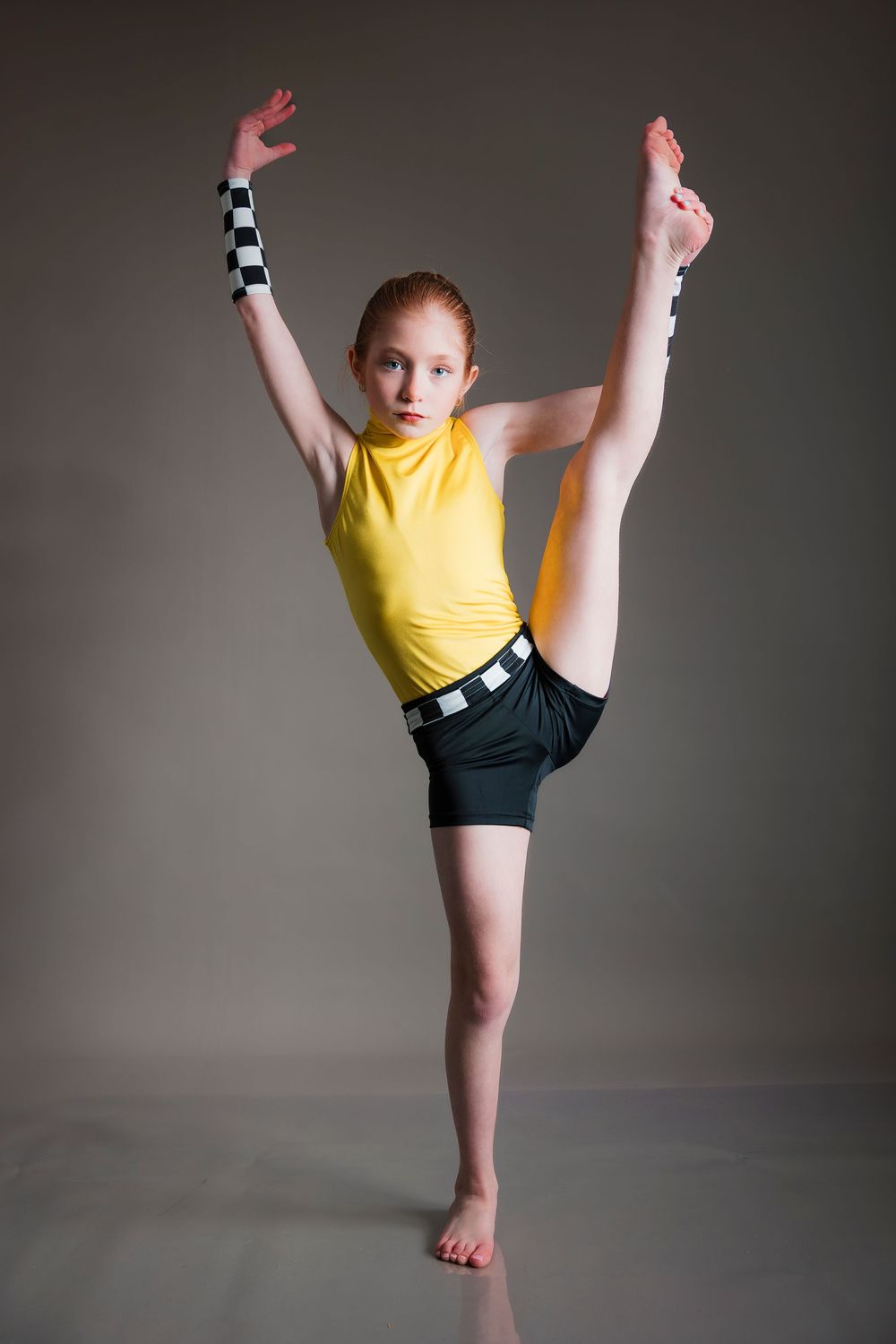 Young dancer in yellow top and black shorts performs a high kick against gray backdrop while striking a graceful pose.