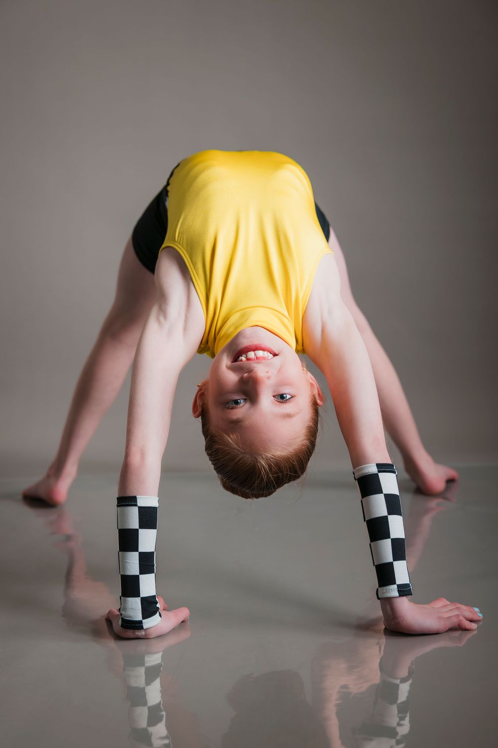 Young person in yellow shirt and checkerboard leggings doing gymnastic bridge pose on reflective gray surface.