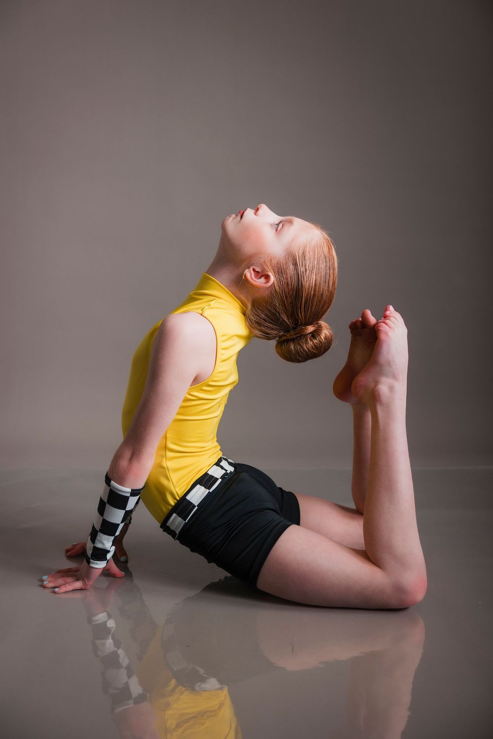 Young dancer in yellow top and black shorts striking a graceful pose on reflective floor.