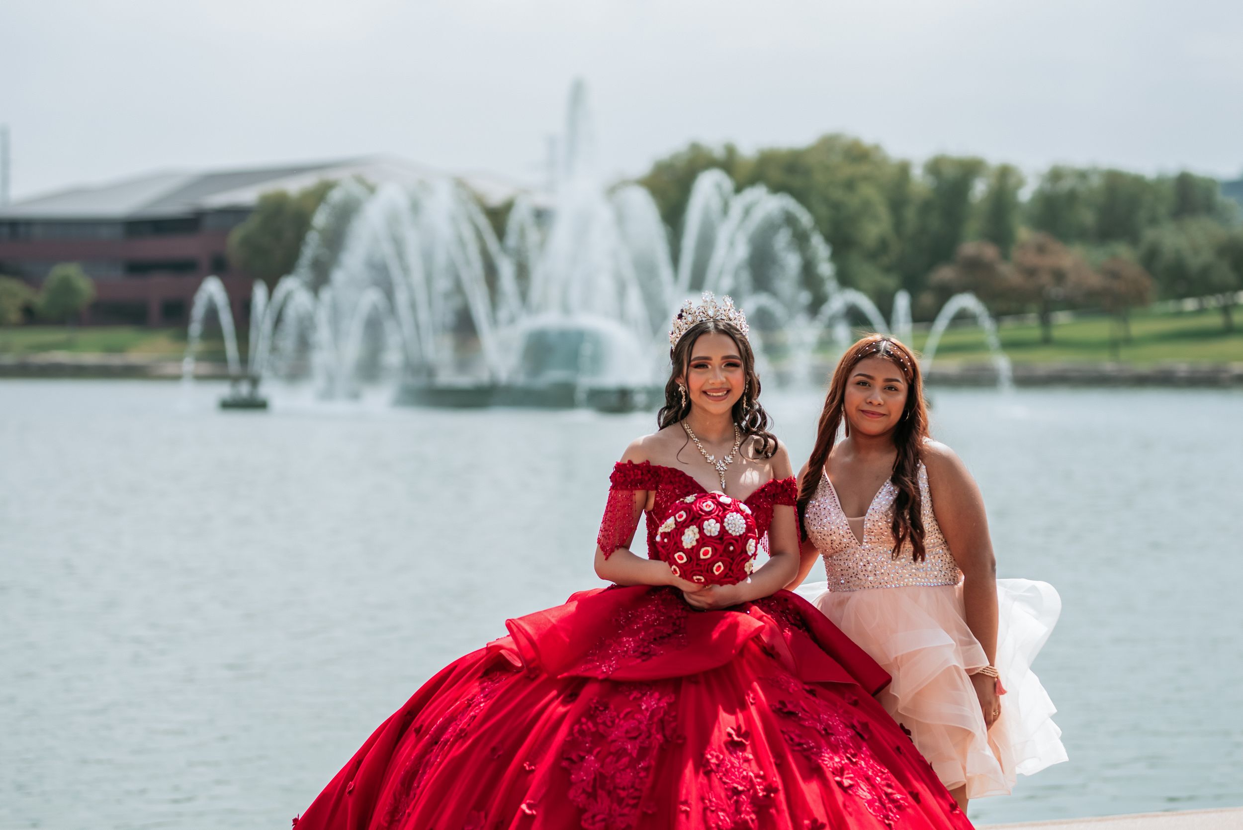 Group photo session by a lake with fountains featuring formal dresses in red and light pink.