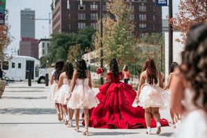 Line of attendants in light colored dresses walking outdoors with cityscape views.