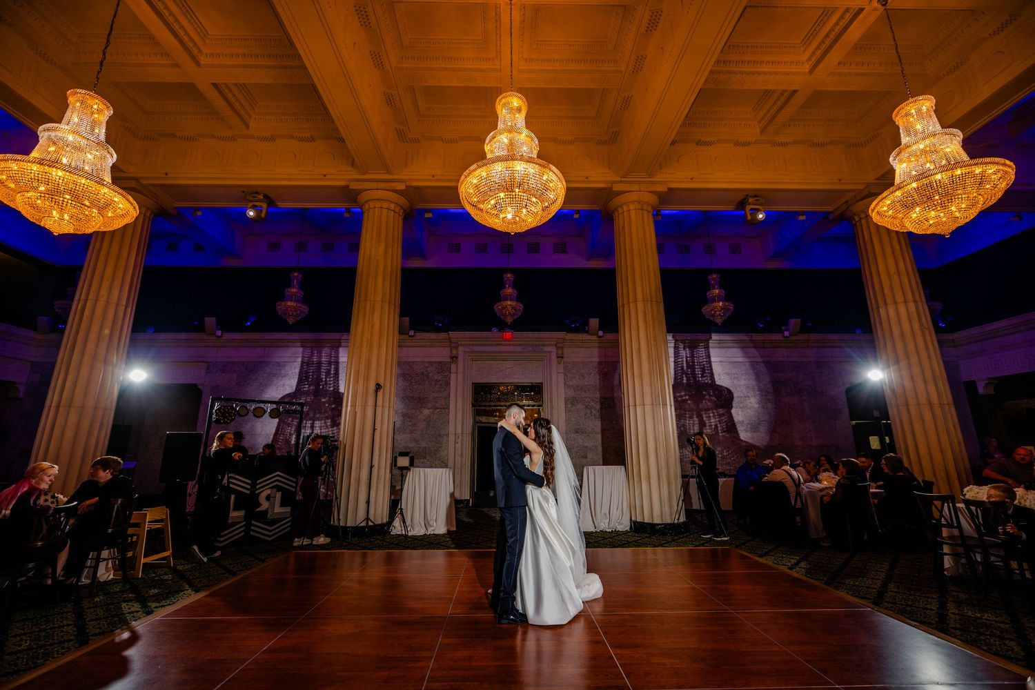 A couple shares their first dance under elegant chandeliers in a grand ballroom with wooden columns and blue lighting Grand Rapids Michigan